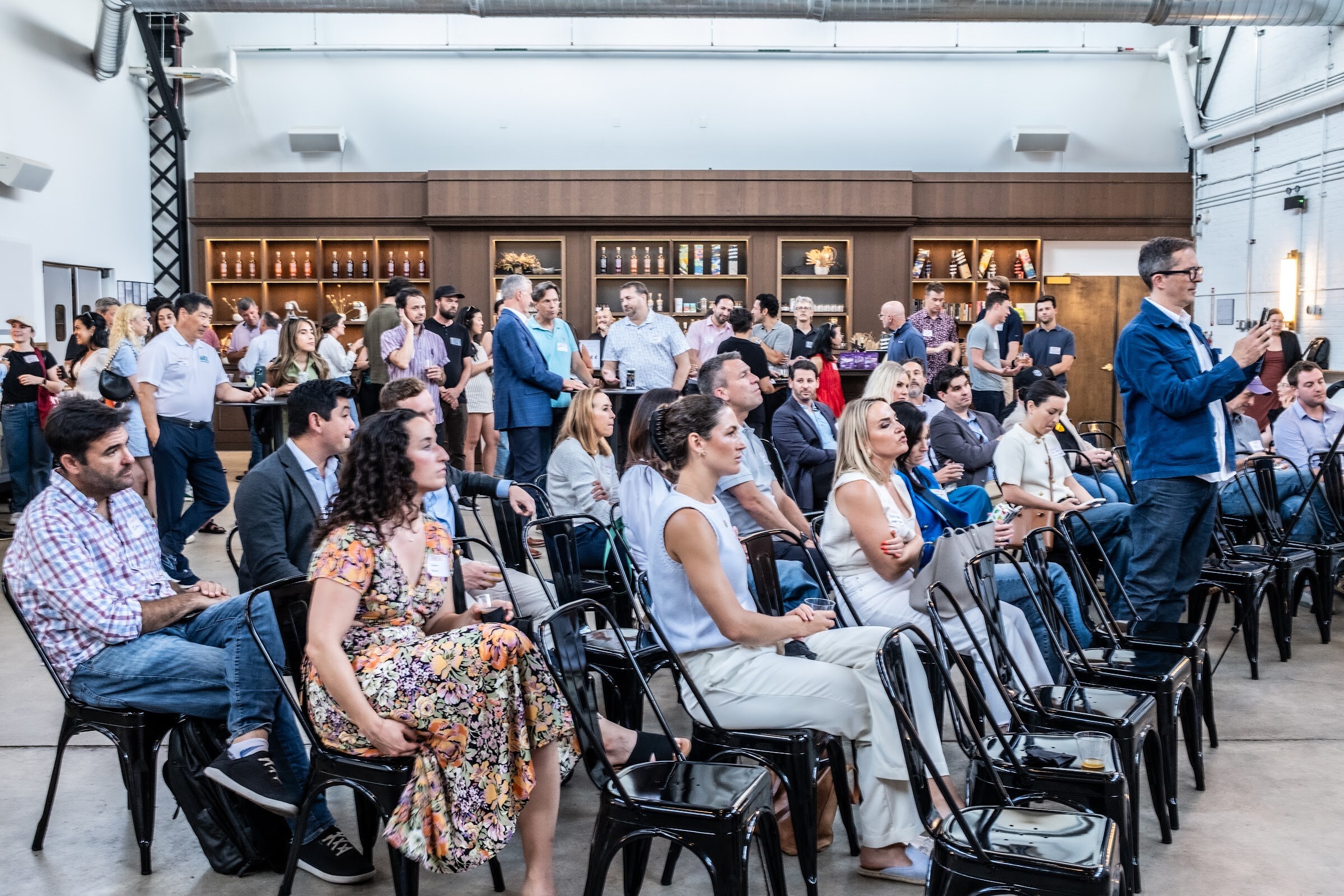 Seated guests in black chairs facing the stage, with a wooden back bar behind them.