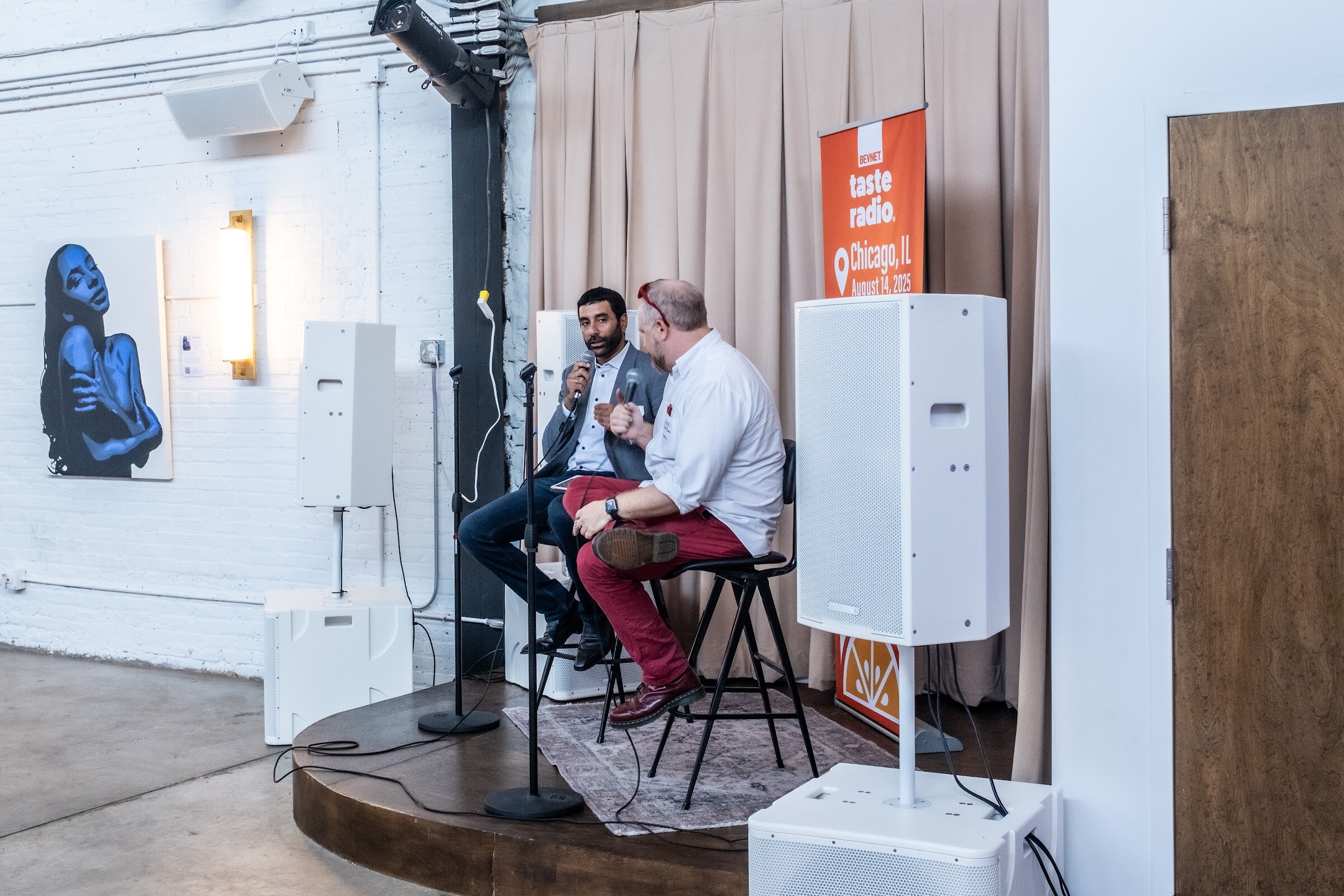 Two men seated on a small stage next to a speaker, with a painting on the wall behind.