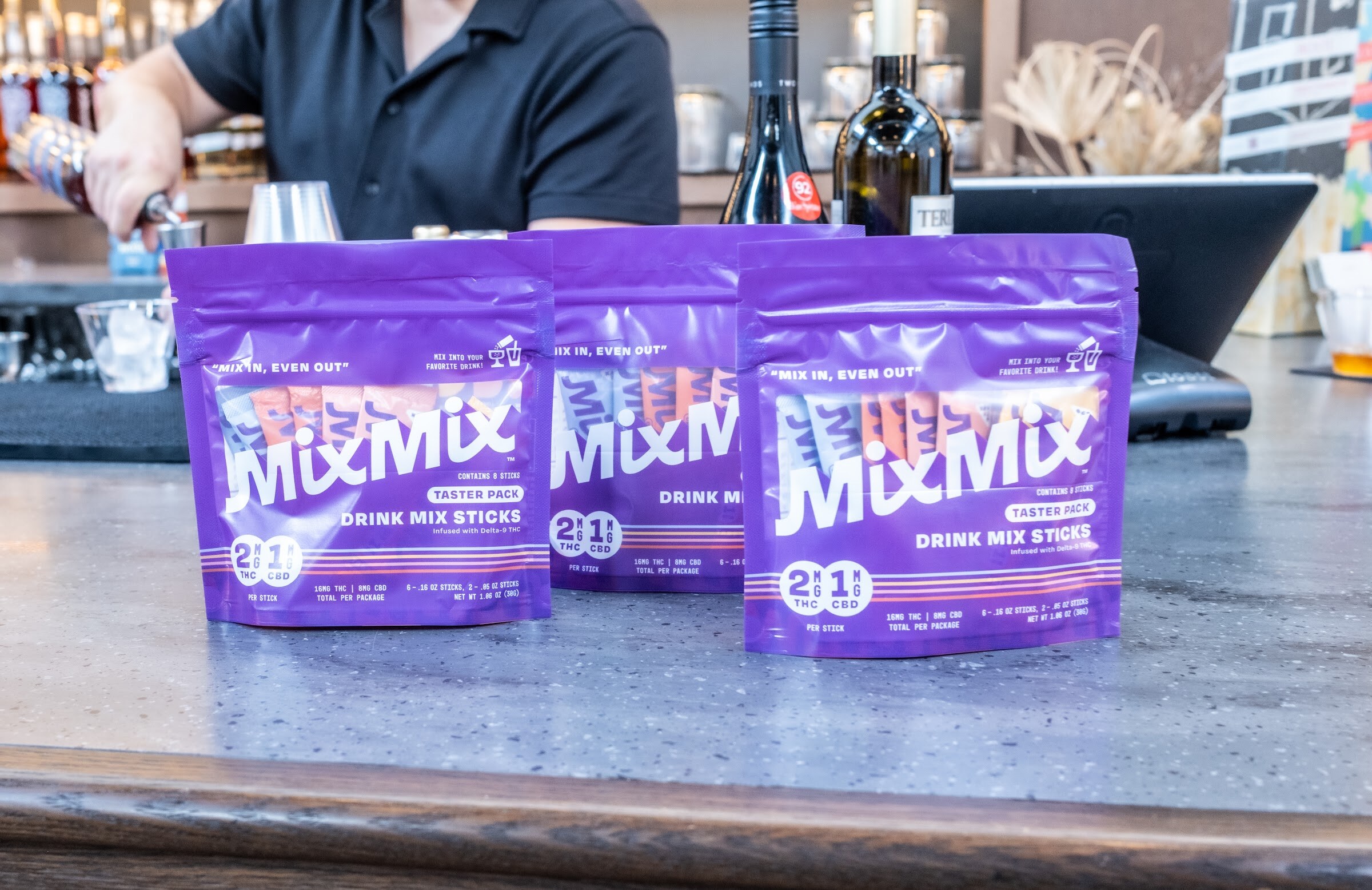 Three purple MixMix drink mix pouches on a bar top with a bartender pouring behind.
