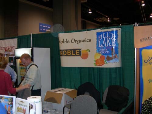 A banner reading Noble Organics and Blue Lake Citrus Products hangs on a green booth wall. A man with a shoulder bag stands near a small refrigerator and a woman in red stands at left.