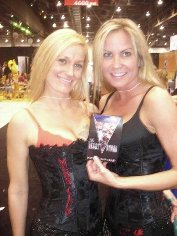 Two smiling blonde women in black lace corsets stand together; one holds up a Las Vegas Vodka box. The corset on the left reads Men's Club.