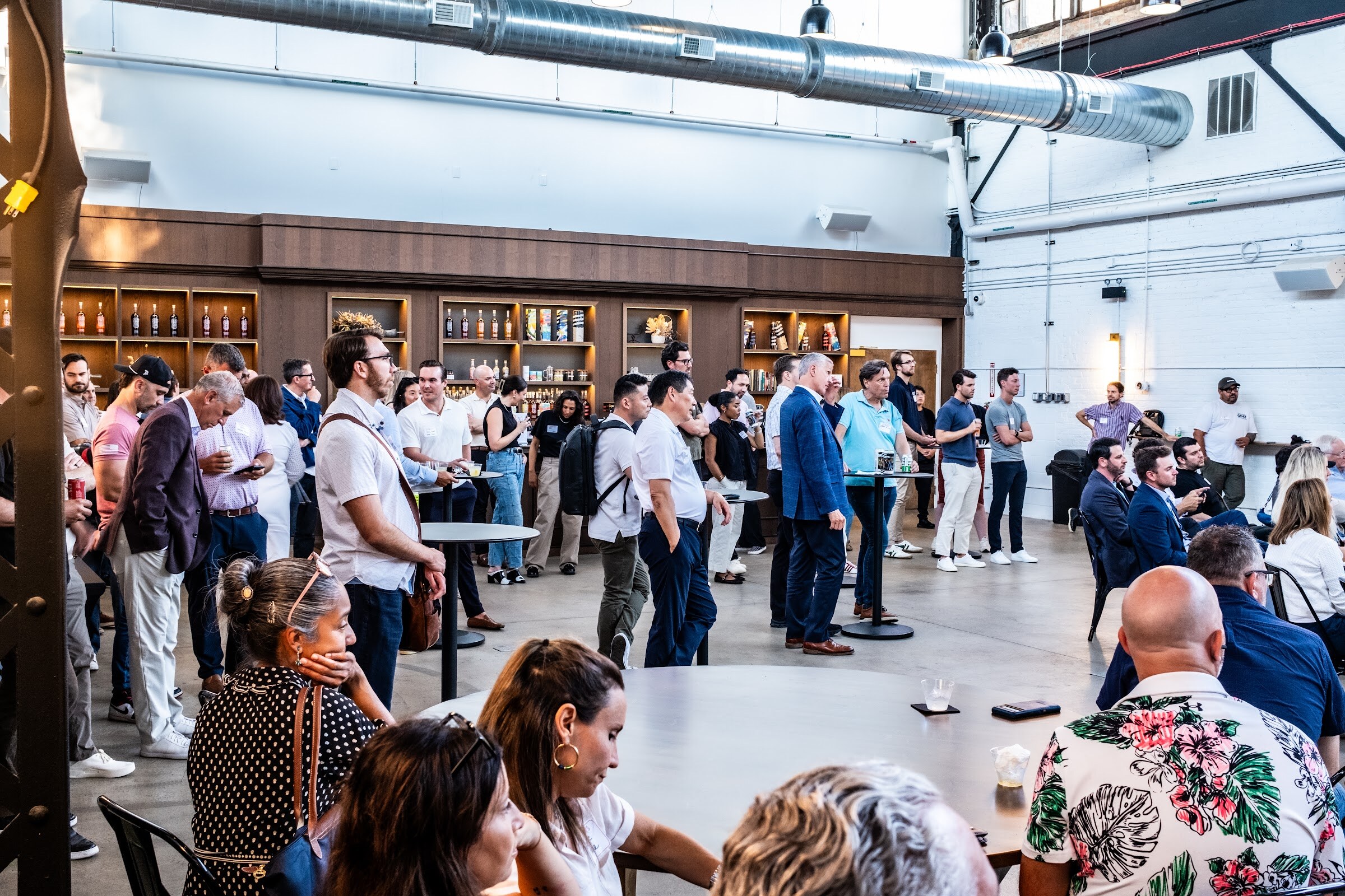 Crowd of standing attendees in front of the room's bar, with seated guests in front.