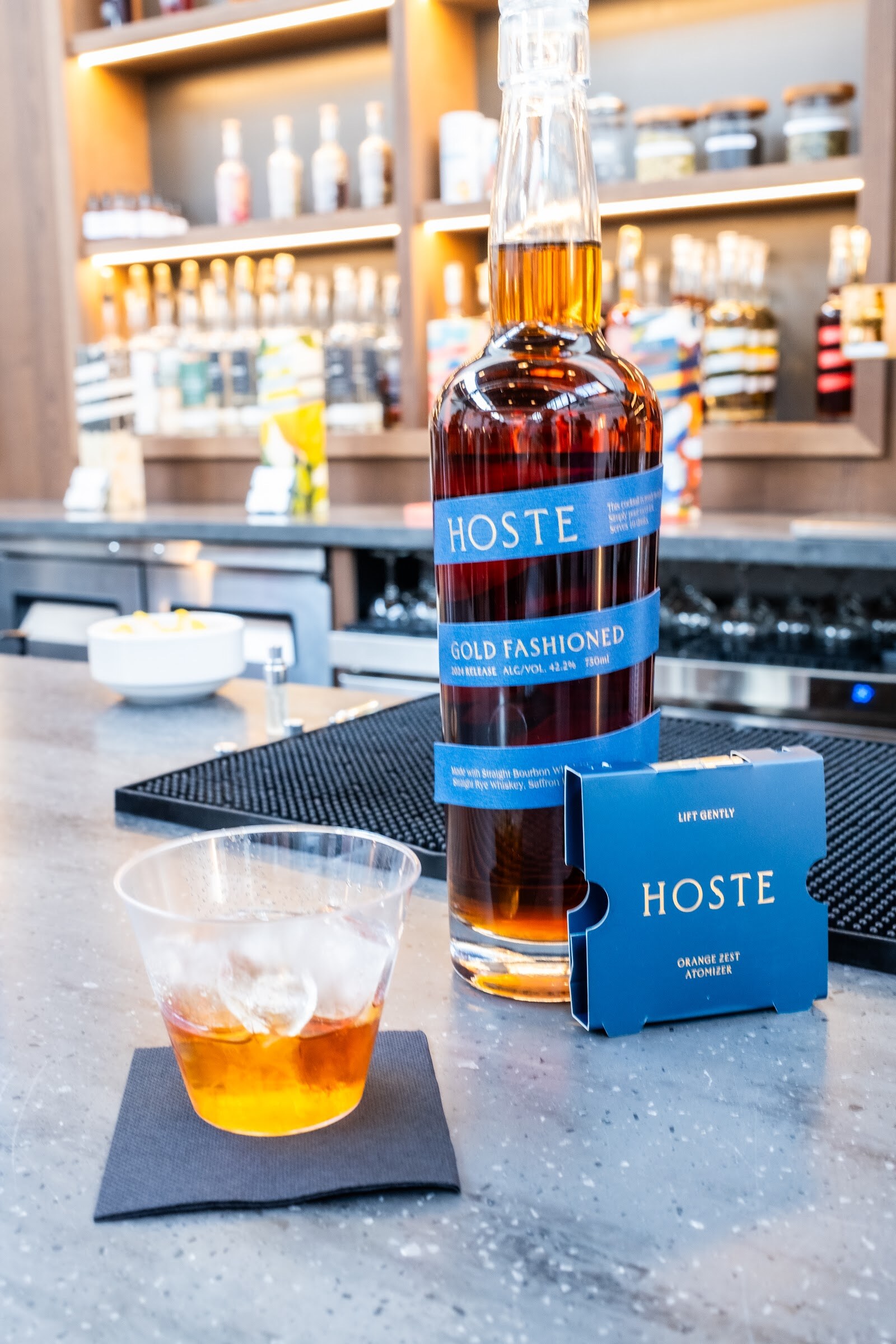 Bottle of Hoste Old Fashioned and a rocks glass on a bar in front of shelves of bottles.