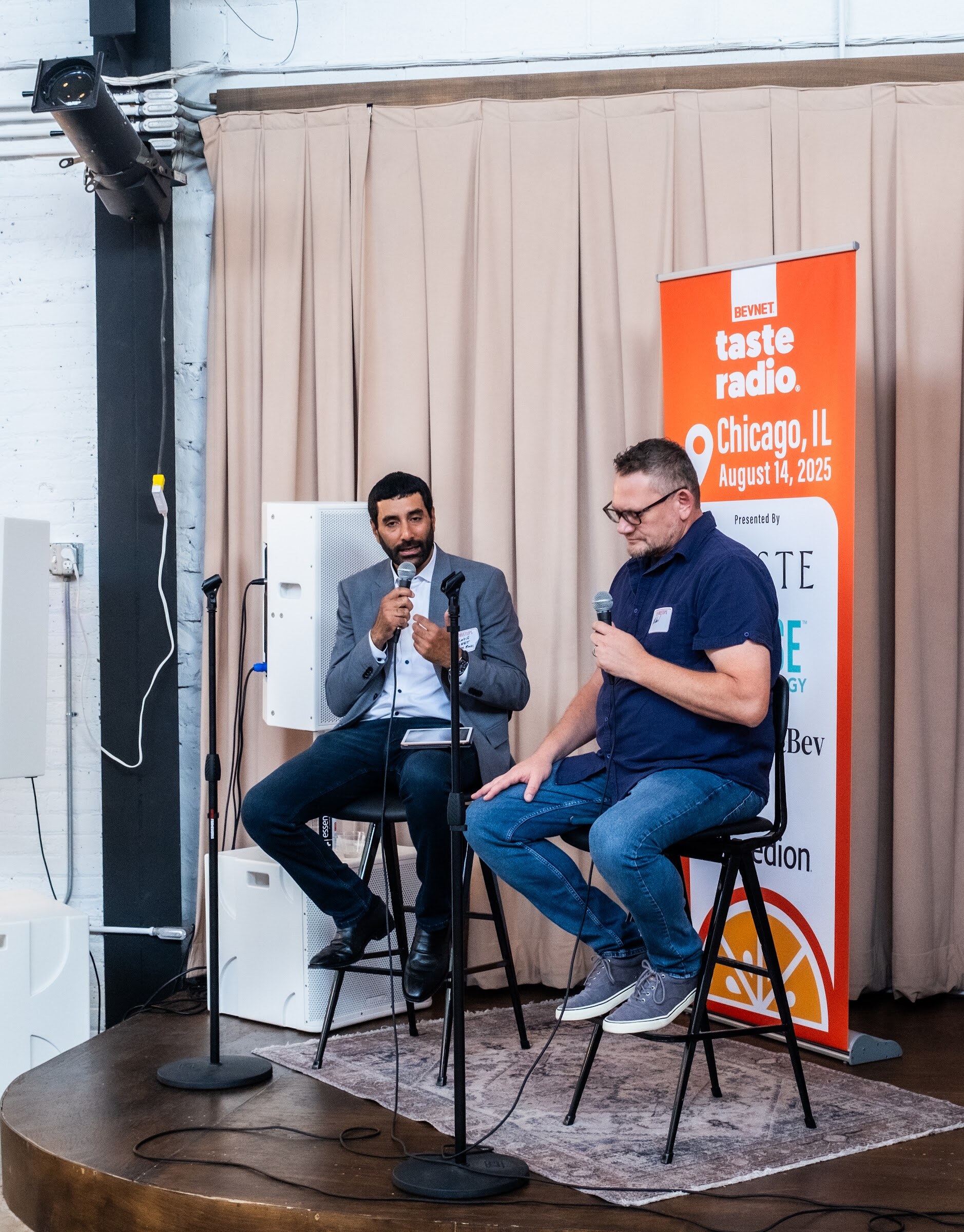 Two men on stools on a small stage in front of the orange Taste Radio banner.