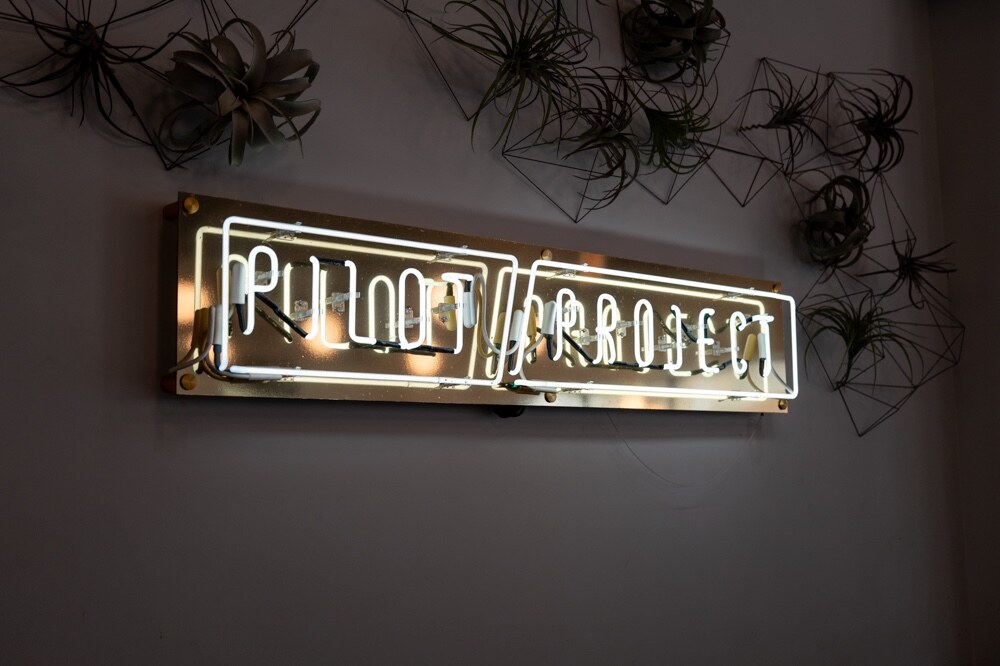 Pilot Project neon sign on a dark wall surrounded by air plants in geometric holders.