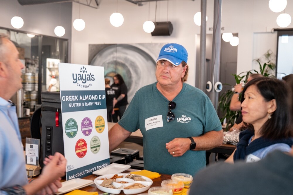 Man in a Yaya’s cap and t-shirt talks with attendees beside a Yaya’s Greek Almond Dip sign and a platter of samples.