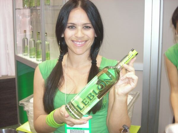 A smiling woman with dark braided pigtails and a green top holds a bottle of Leblon cachaça. Glass shelves with more bottles stand behind her.
