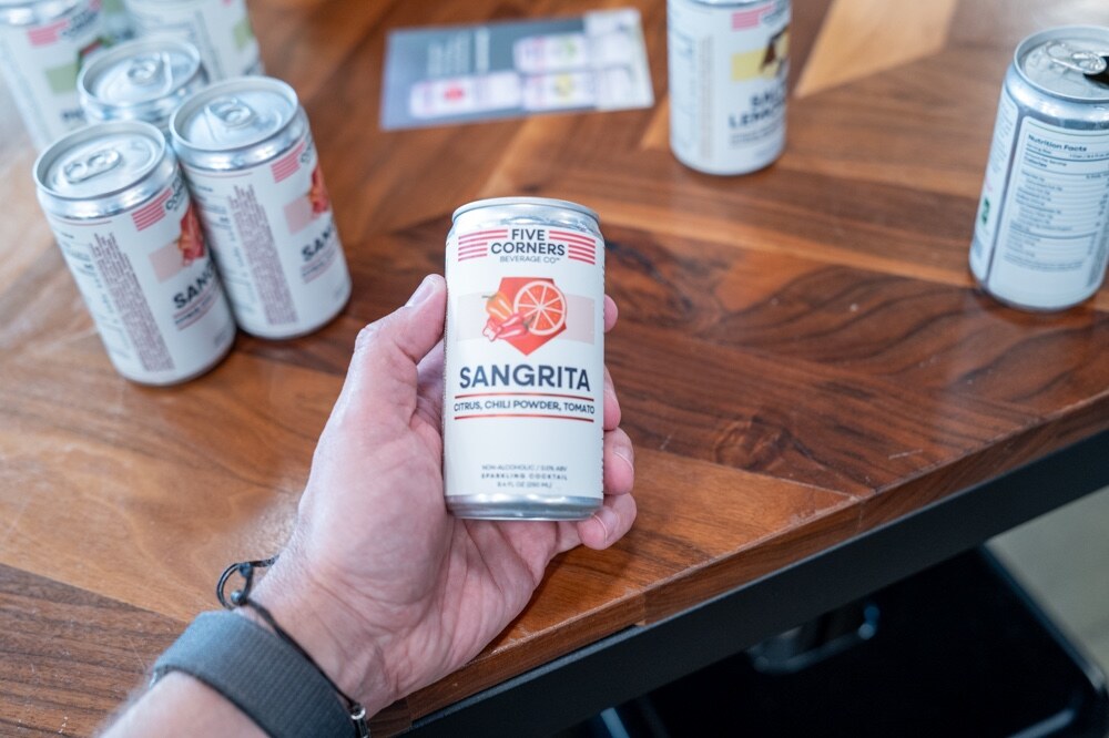 Hand holding a small can of Five Corners Sangrita over a wooden table with more cans.