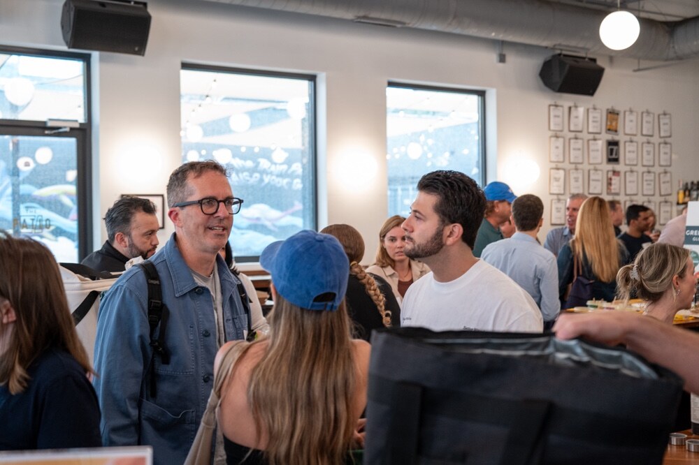 Smiling man in glasses and a denim jacket talks with attendees in a busy taproom.