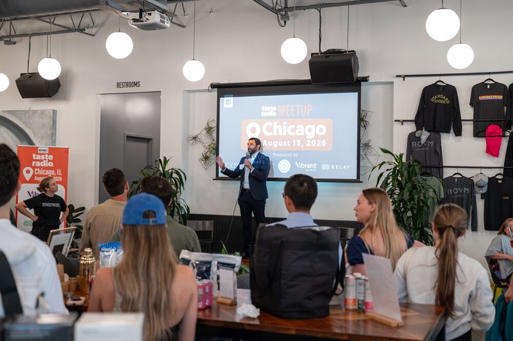Host in a navy suit speaks into a microphone beside a screen reading Taste Radio Meetup Chicago August 13, 2026, with attendees seated in the foreground.