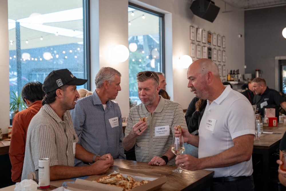 Four men stand around a table with a pizza box, one holding a wine glass.