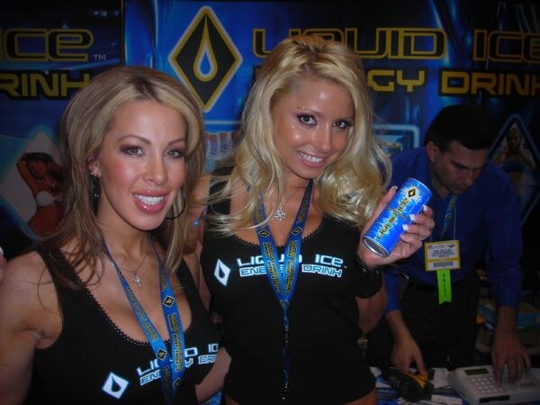 Two smiling women in black Liquid Ice Energy Drink tops and lanyards pose at the booth; one holds a can. A man in a blue shirt stands behind them.