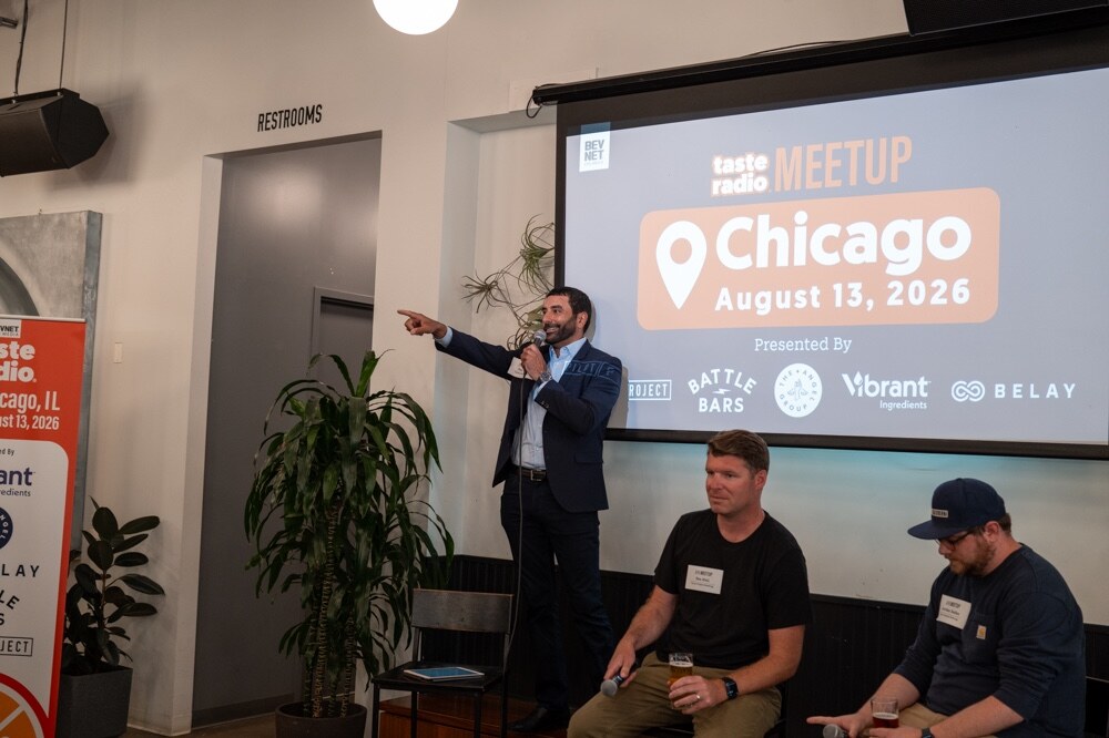 Host in a suit points while two seated guests in black t-shirts wait beneath a screen reading Taste Radio Meetup Chicago.