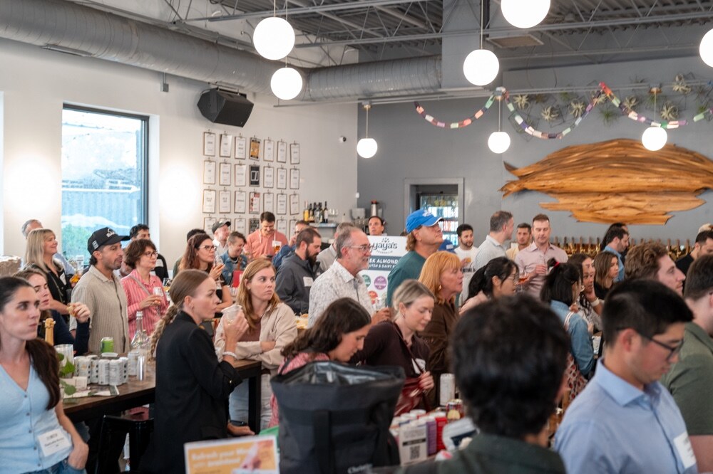 Crowd seated and standing around long tables in a taproom, facing the stage.