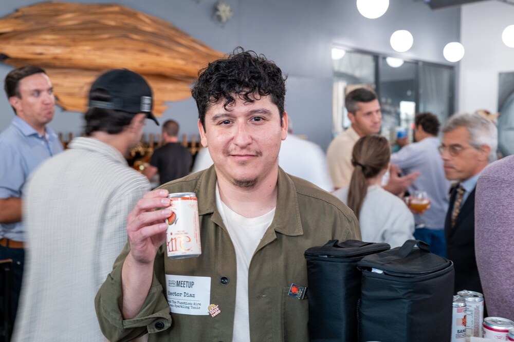 Man in an olive jacket smiles holding a small can of Aire in a crowded taproom.