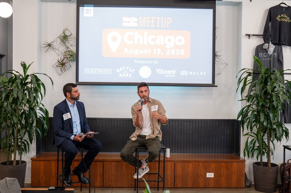Host in a navy suit and a guest in a tan jacket sit on stools on a low stage in front of the Chicago meetup screen and plants.