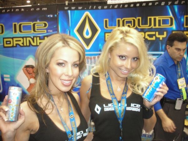 Two women in black Liquid Ice Energy Drink tops each hold up a can in front of a blue Liquid Ice banner. A man in a blue shirt stands behind them.