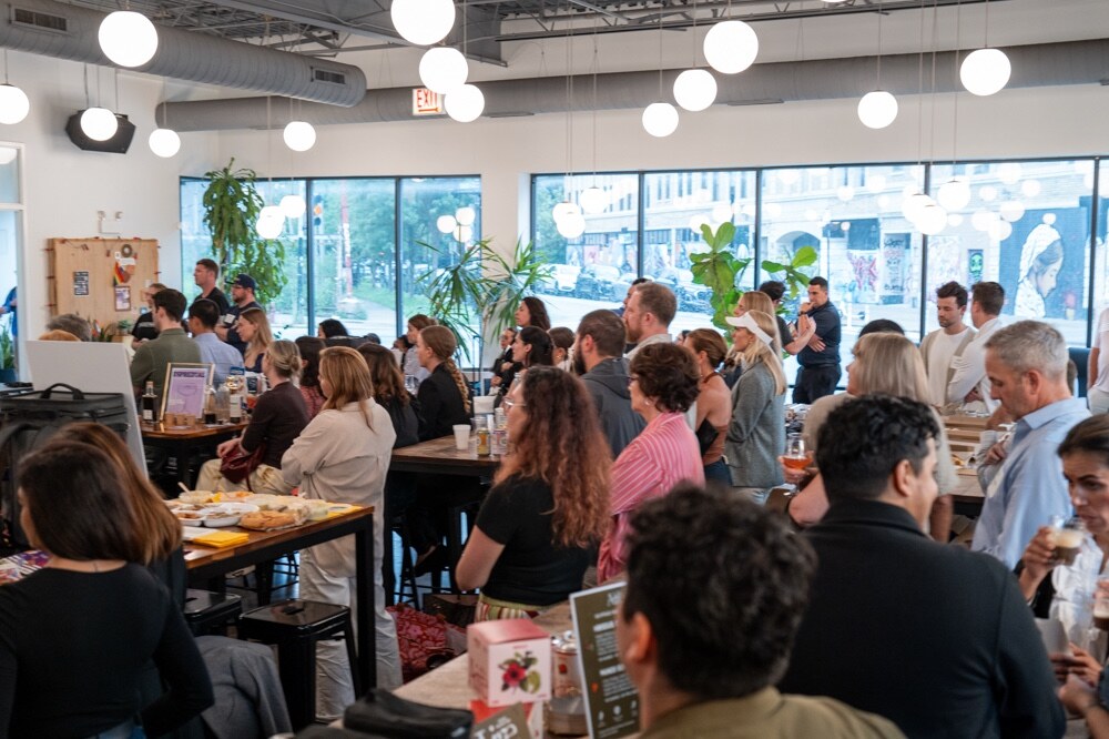 Crowd seated at tables in a taproom with large windows and hanging globe lights.