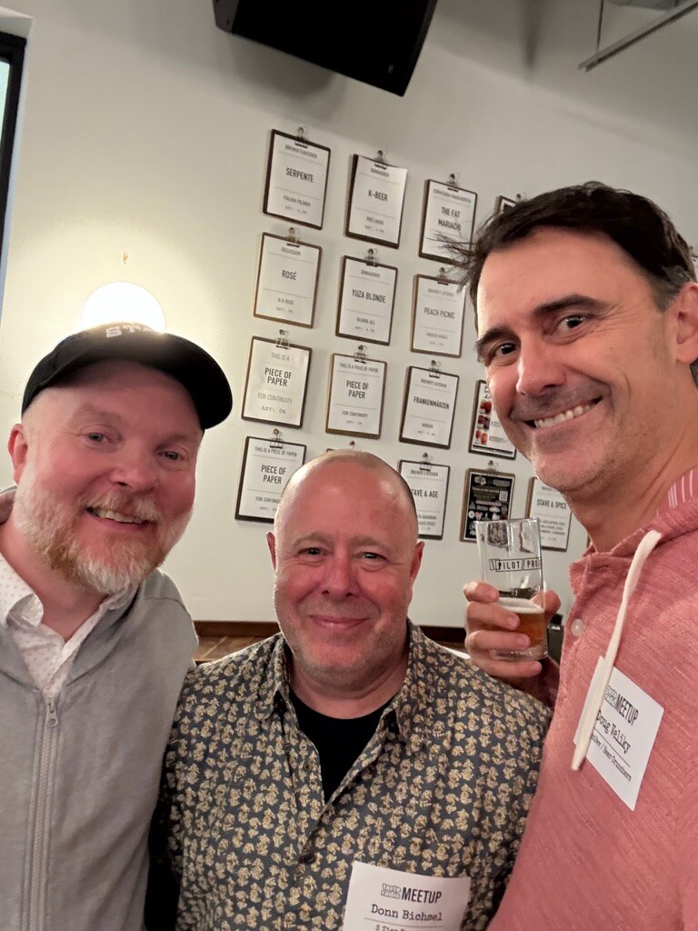 Three smiling men take a selfie in front of a wall of framed beer menus, one holding a glass of beer.