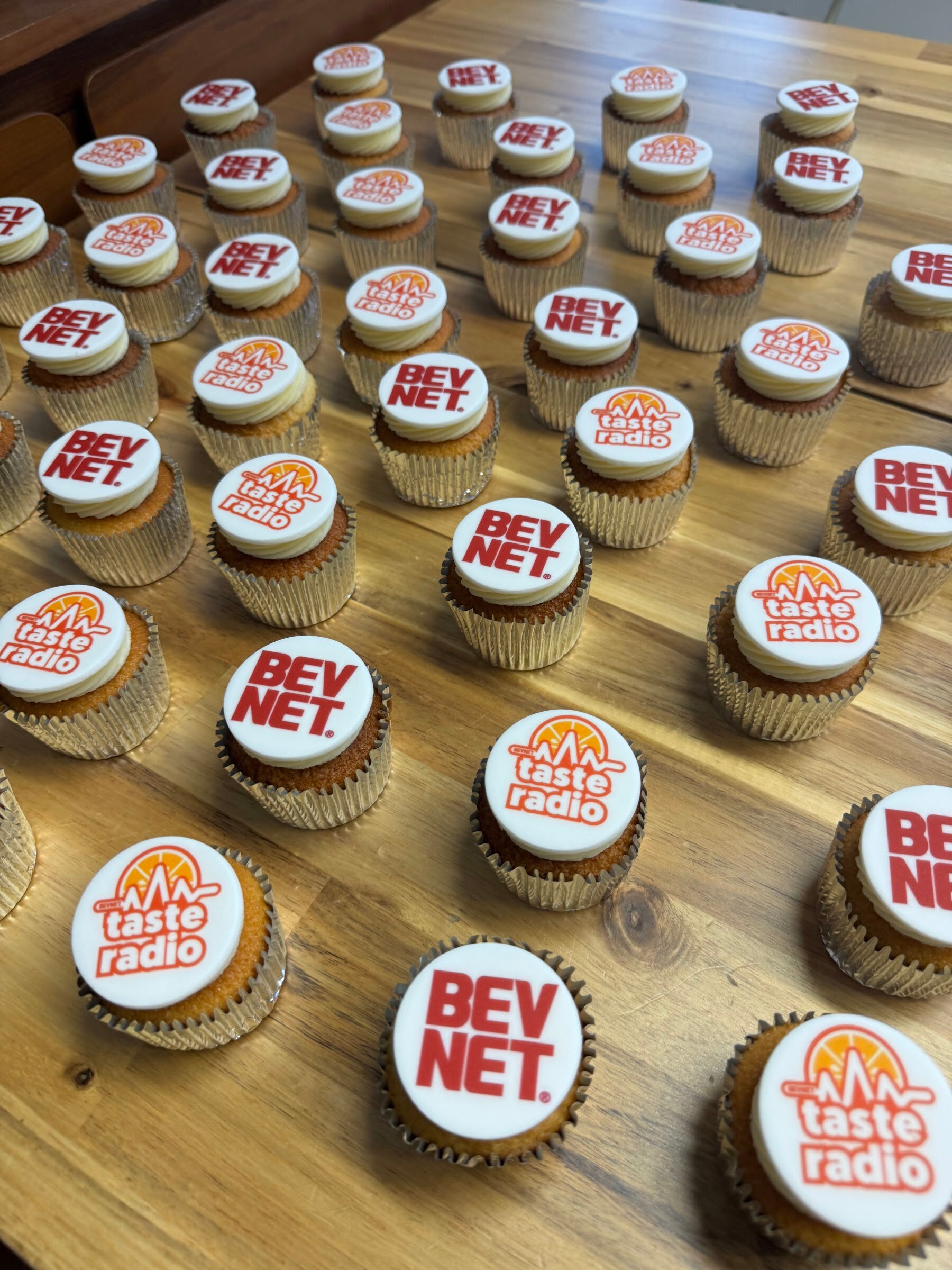Wooden table covered in cupcakes decorated with BevNET and Taste Radio logos.