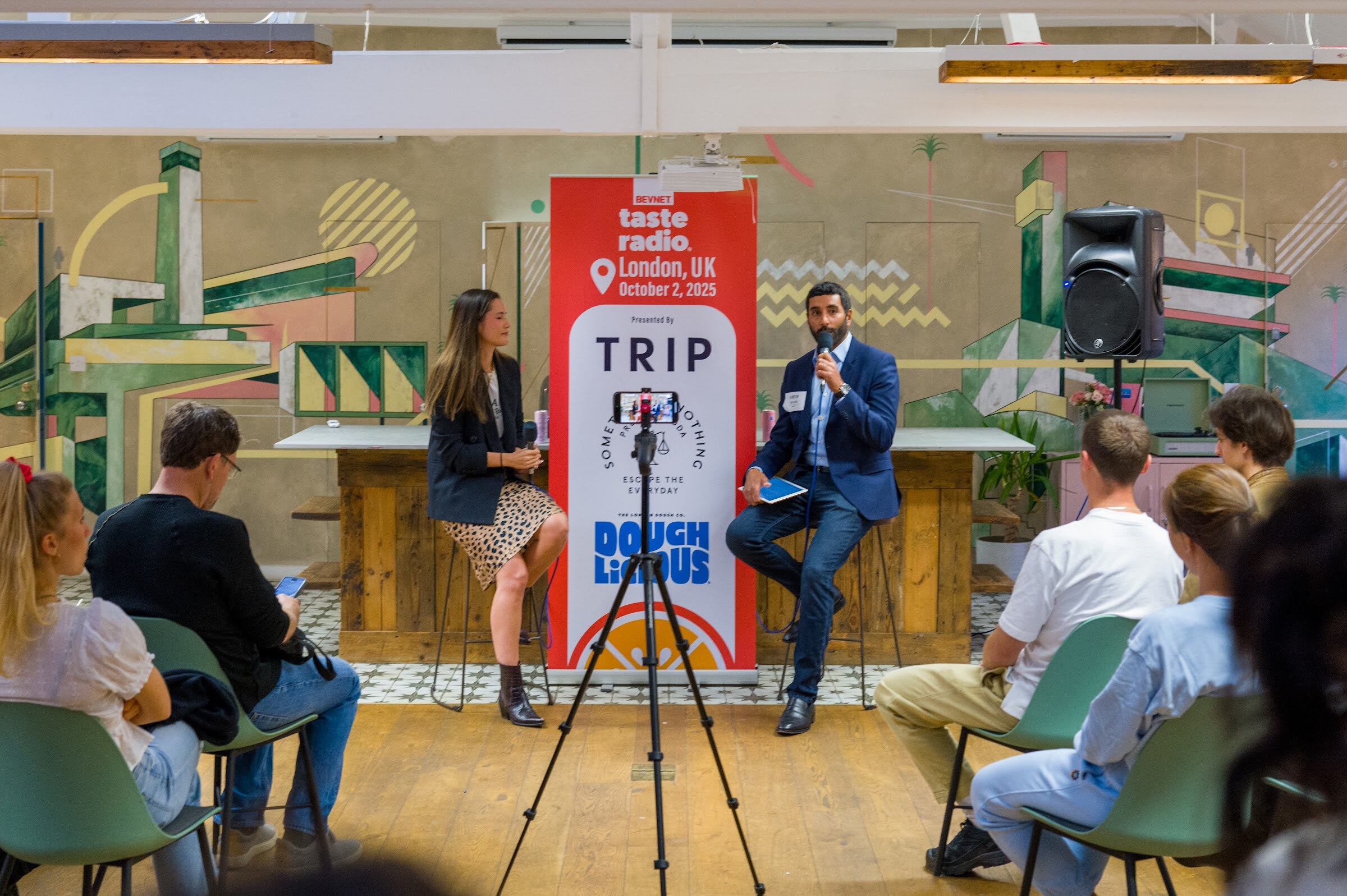 Woman in a dark blazer and man in a blue suit seated on stools in front of the banner, with an audience at right.