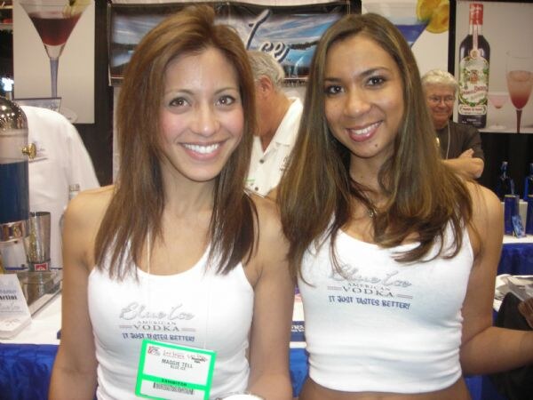 Maggie Tell, on the left, and another woman smile in white Blue Ice American Vodka tank tops at a booth with cocktail glasses and a bottle in the background.