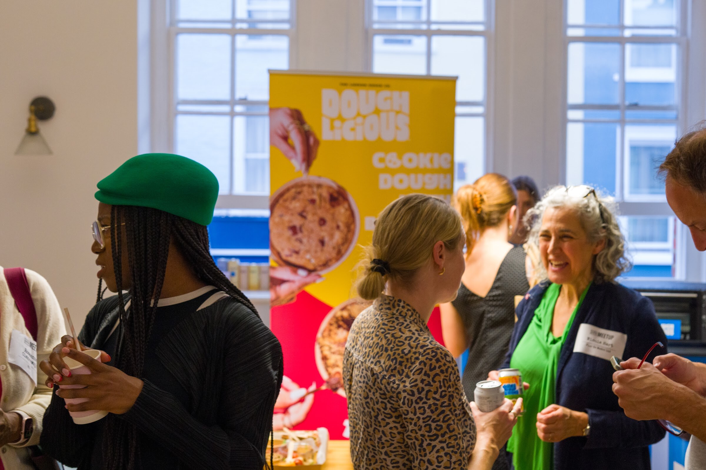 Attendees chatting in front of a yellow and red Doughlicious banner by a window.
