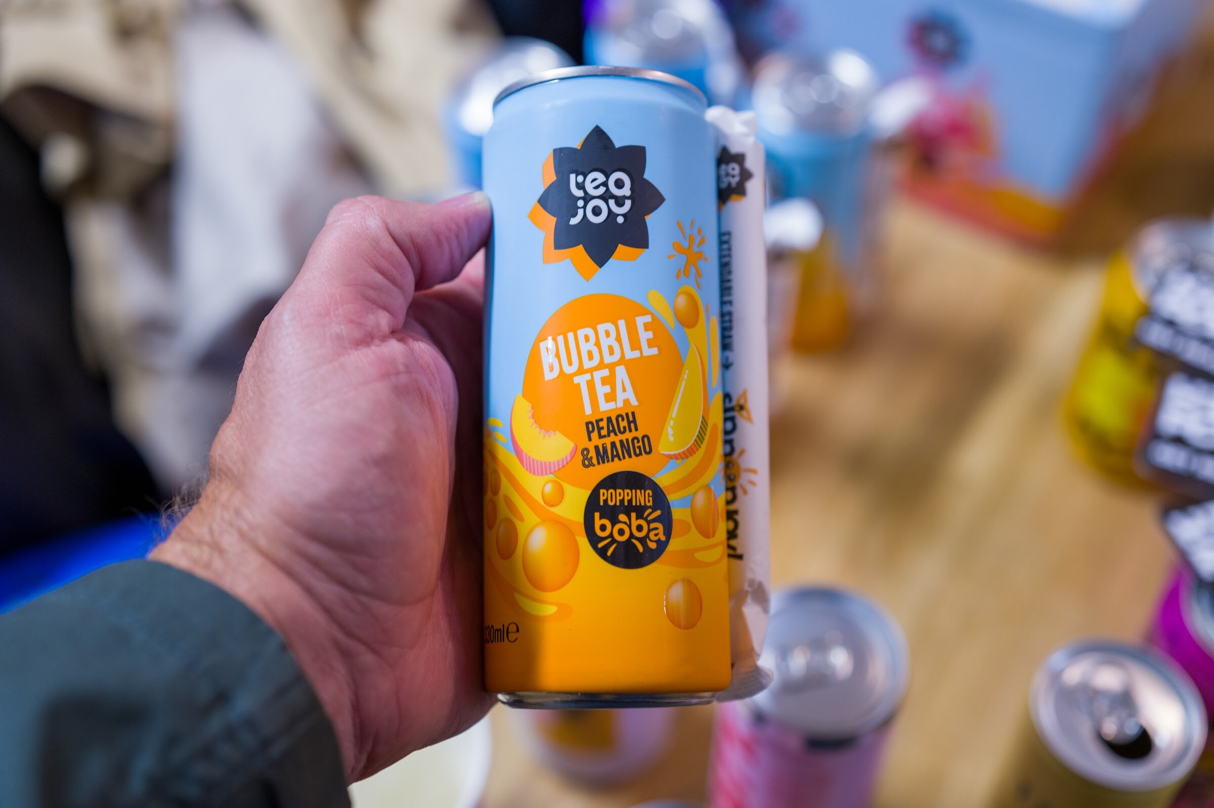 Hand holding a can of Tea Joy bubble tea against a blurred table of cans.