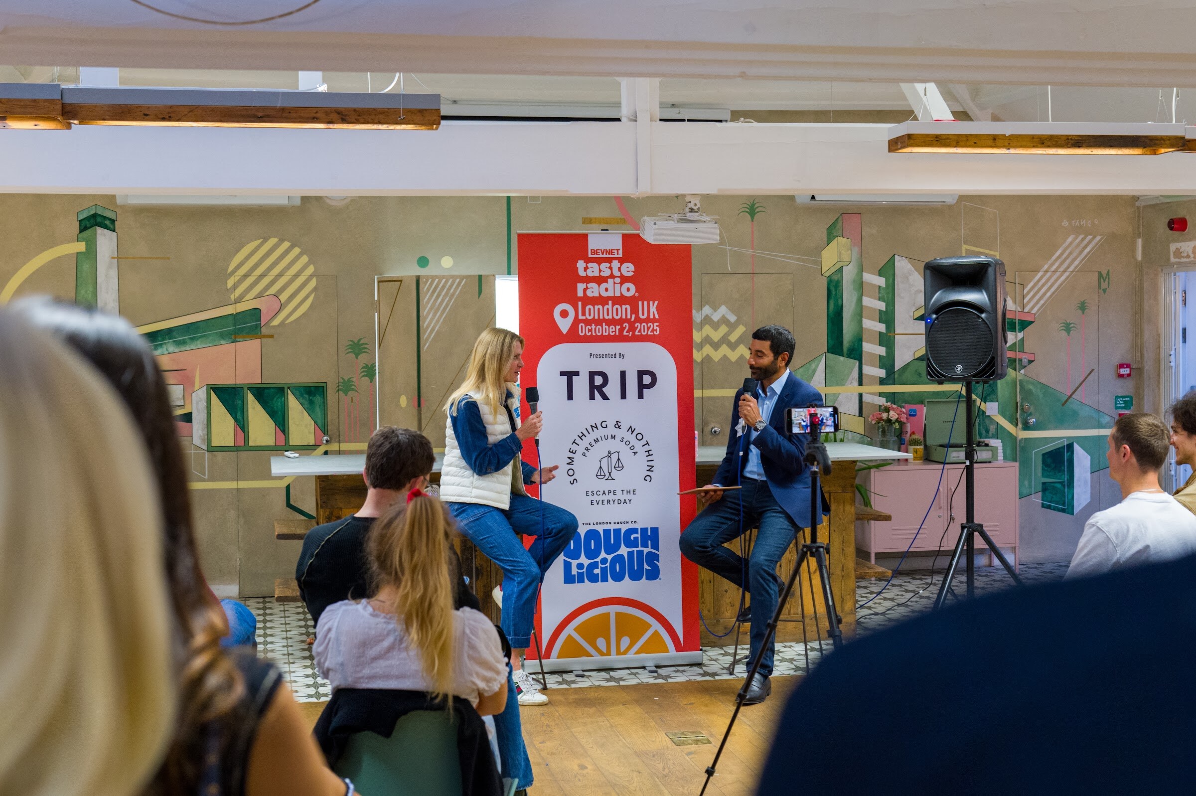 Woman in a white vest and man in a blue suit holding microphones on stools in front of the banner.