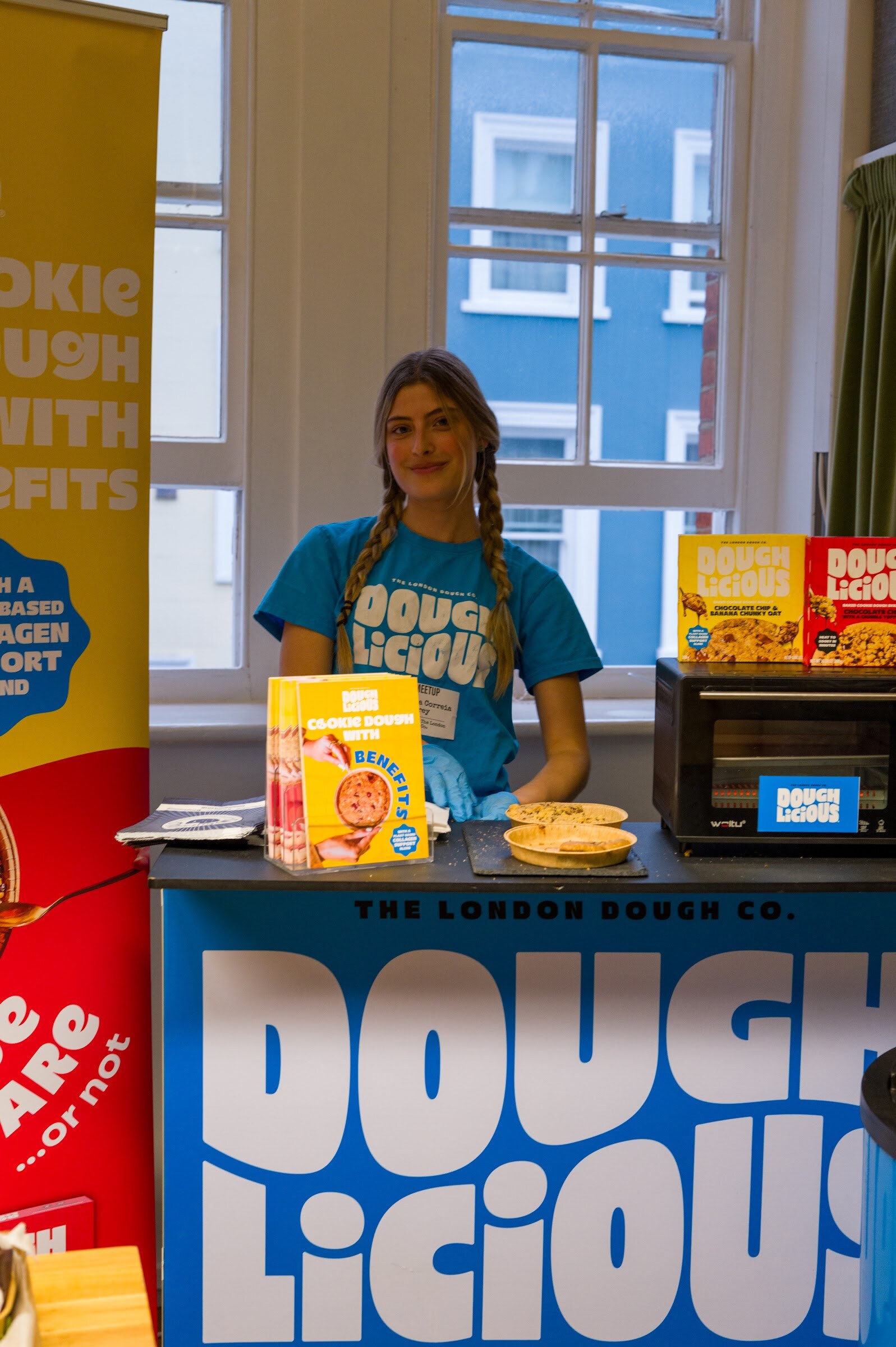 Smiling woman in a Doughlicious shirt behind a table with cookie dough boxes and a toaster oven.