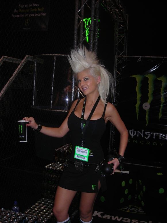 A smiling woman with spiked platinum hair and a black outfit holds a Monster Energy cup in a dark booth with Monster logos and stacked cans.