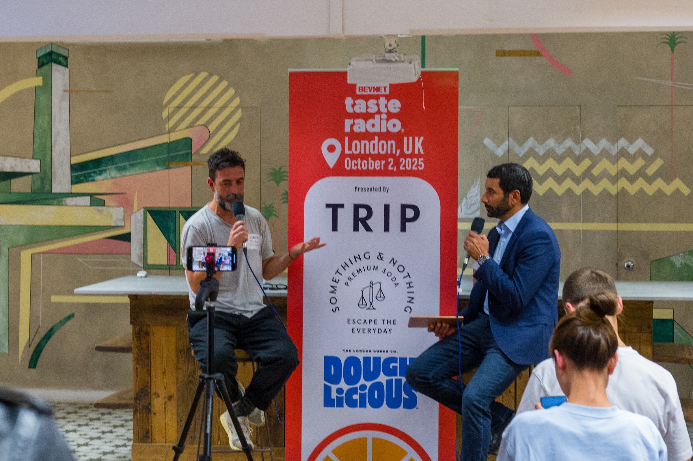 Two men on stools before the Taste Radio London banner, one gesturing as he speaks.