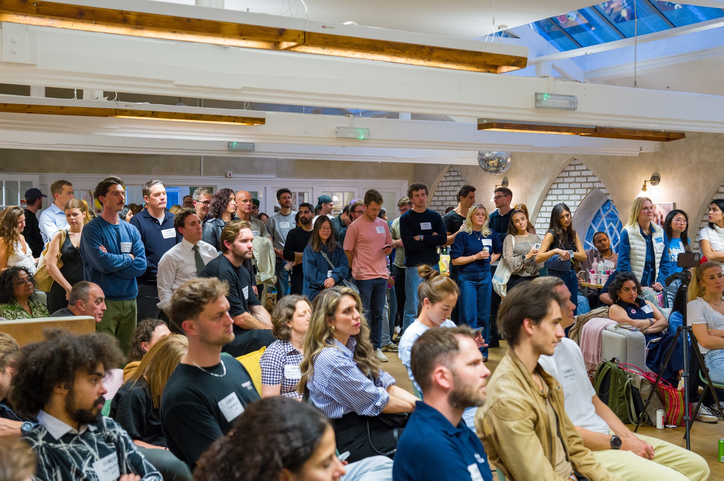 Crowd of seated and standing attendees in a high-ceilinged room with arched windows.