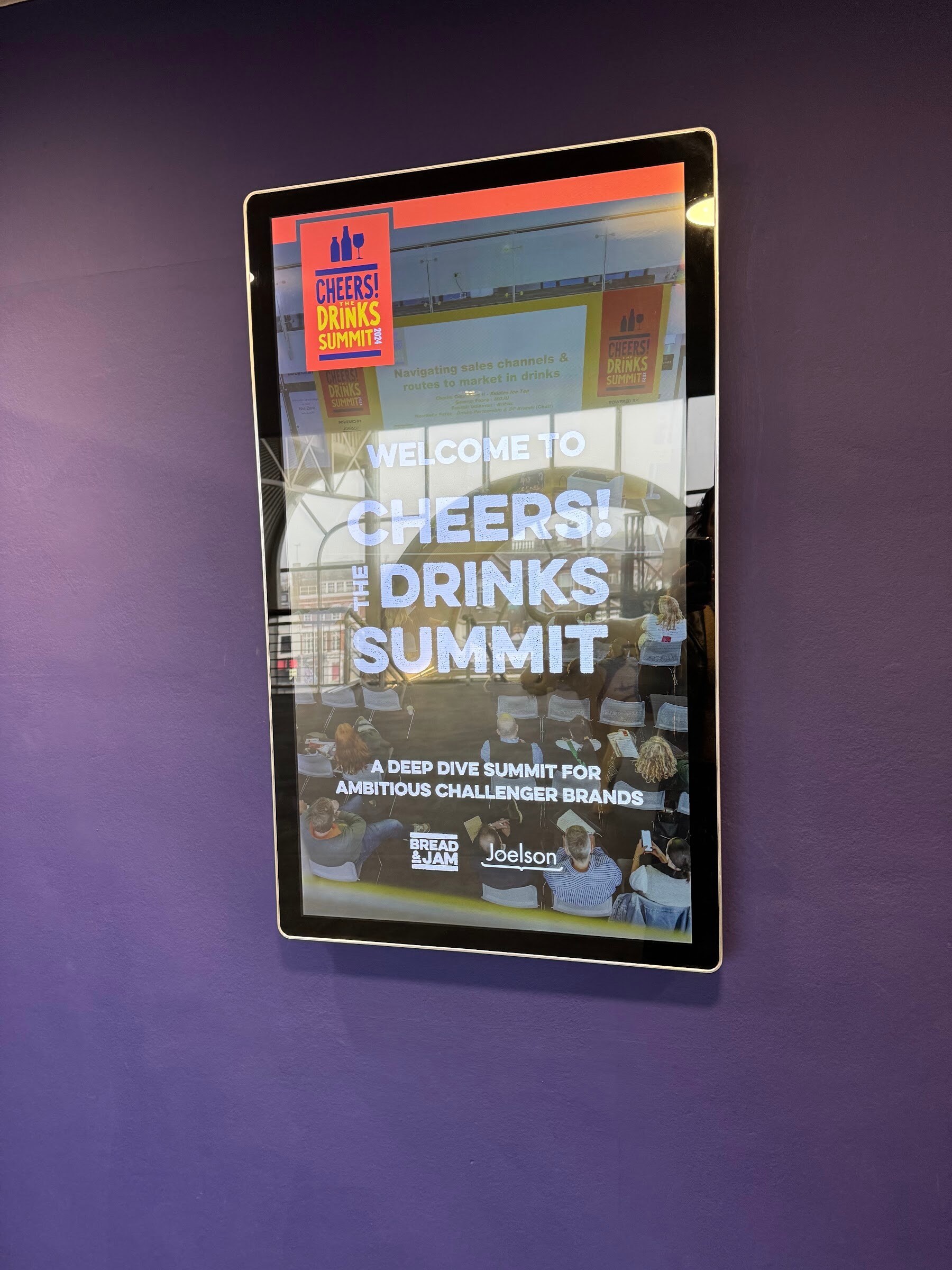 Wall-mounted screen reading Welcome to Cheers! The Drinks Summit, a deep dive summit for ambitious challenger brands.