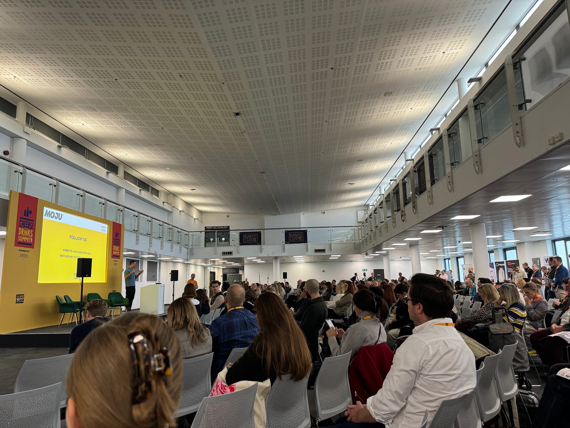 Large hall full of seated attendees facing a stage with a yellow Cheers! The Drinks Summit backdrop.