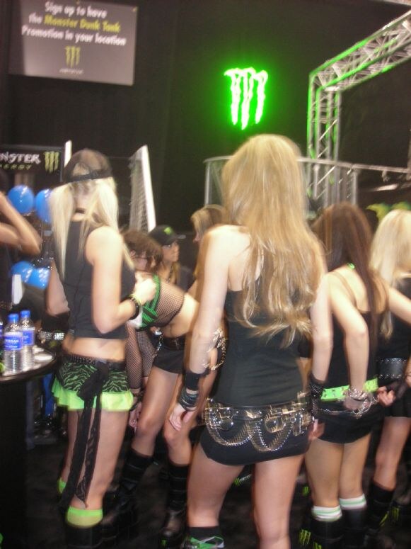 A crowd of women in black and green Monster Energy outfits stands in a booth with a glowing green Monster logo and a sign inviting visitors to sign up for the Monster Dunk Tank promotion.