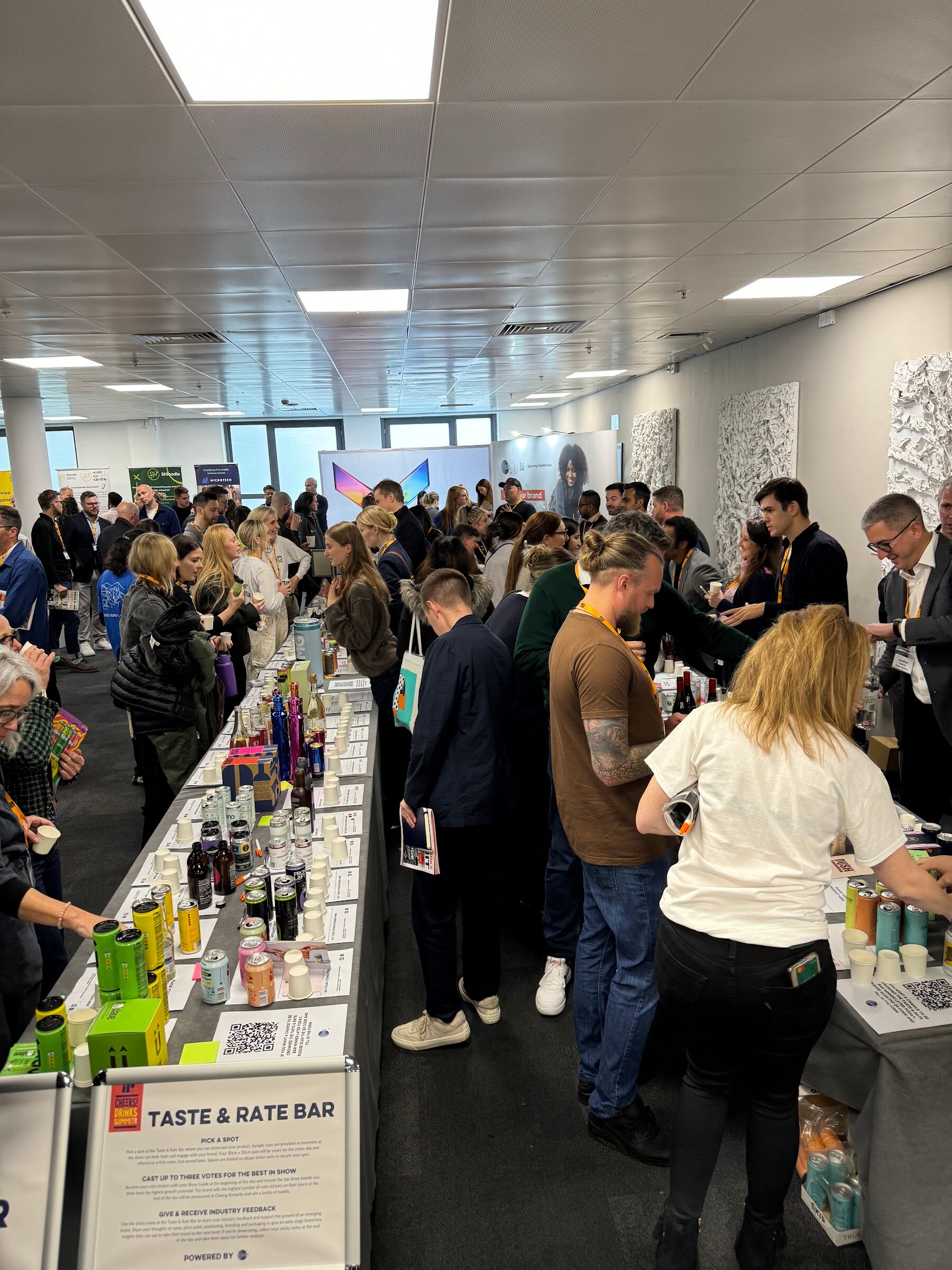 Many attendees along a long table filled with cans and bottles, with Taste and Rate Bar signs in the foreground.