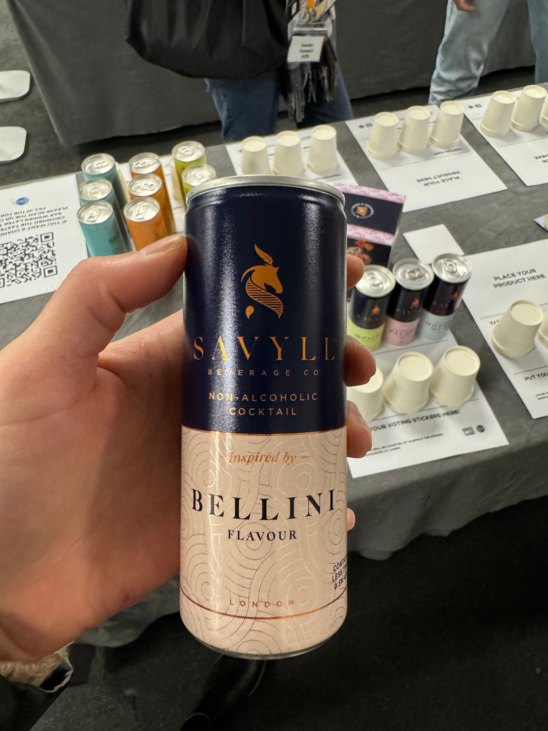 Hand holding a navy and cream can of Savyll non-alcoholic cocktail over a table of tasting cups.