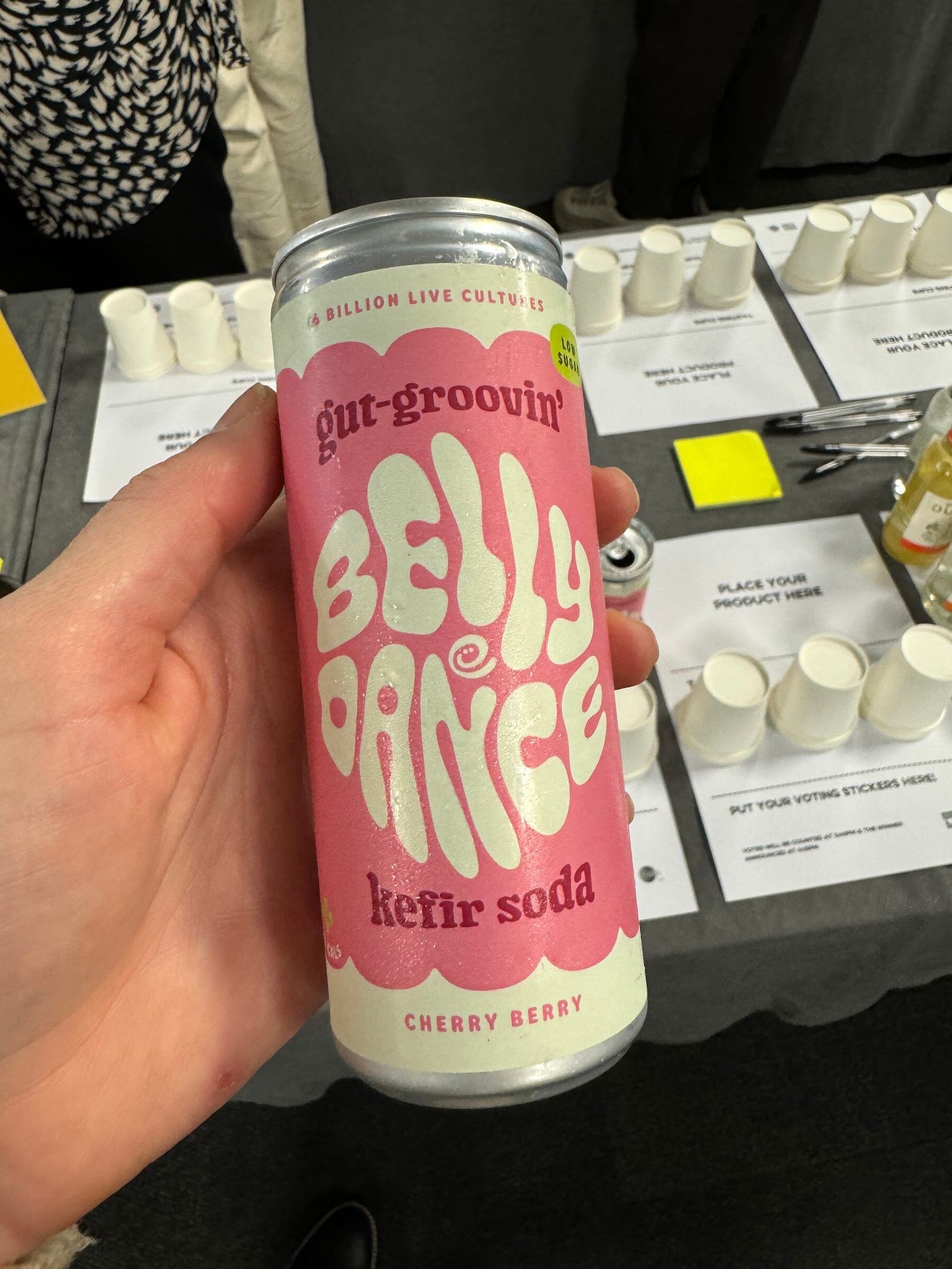 Hand holding a pink can of Belly Dance kefir soda over a table with paper tasting cups.