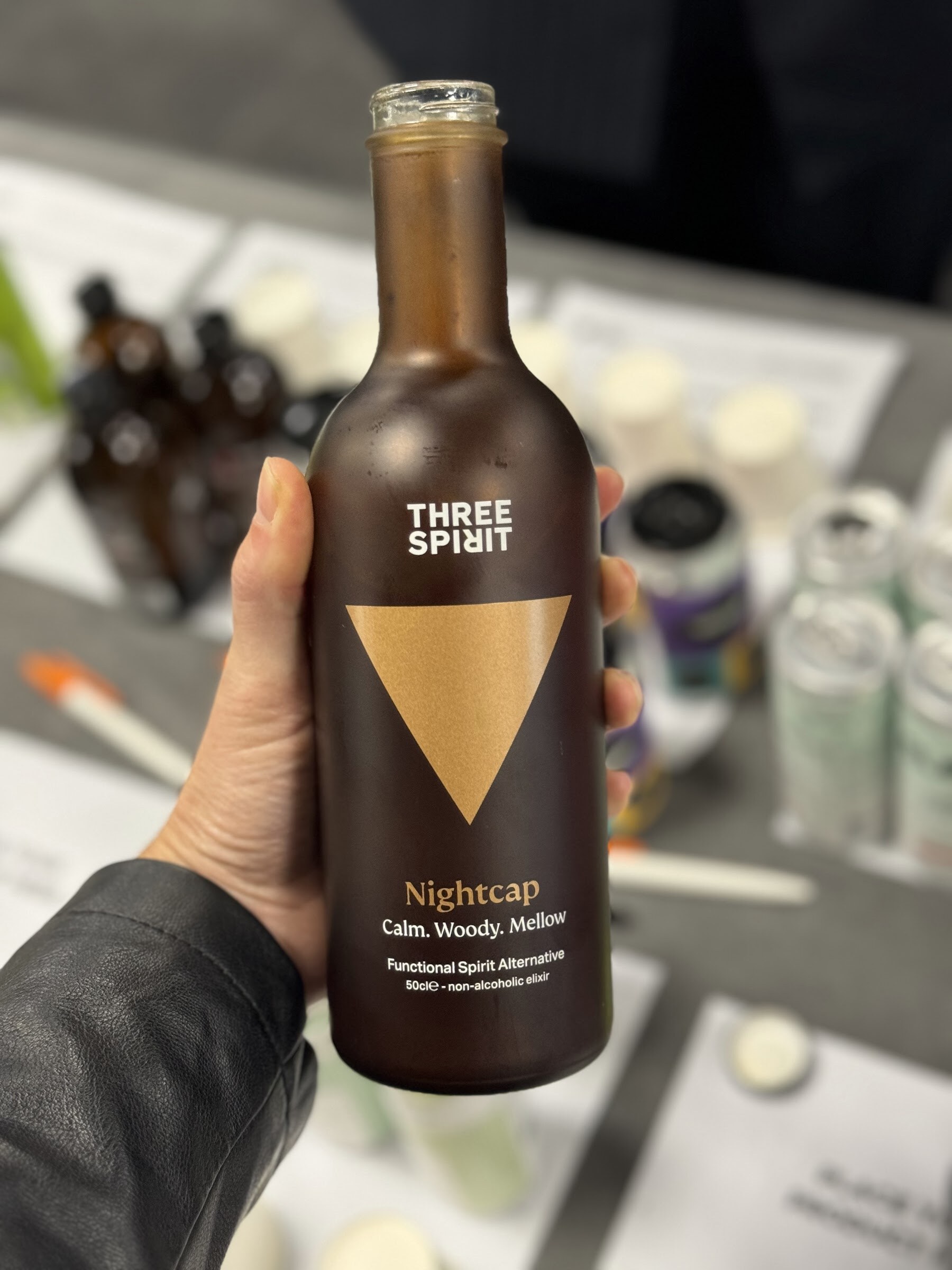 Hand holding a dark bottle of Three Spirit Nightcap above a tasting table with sampling sheets.