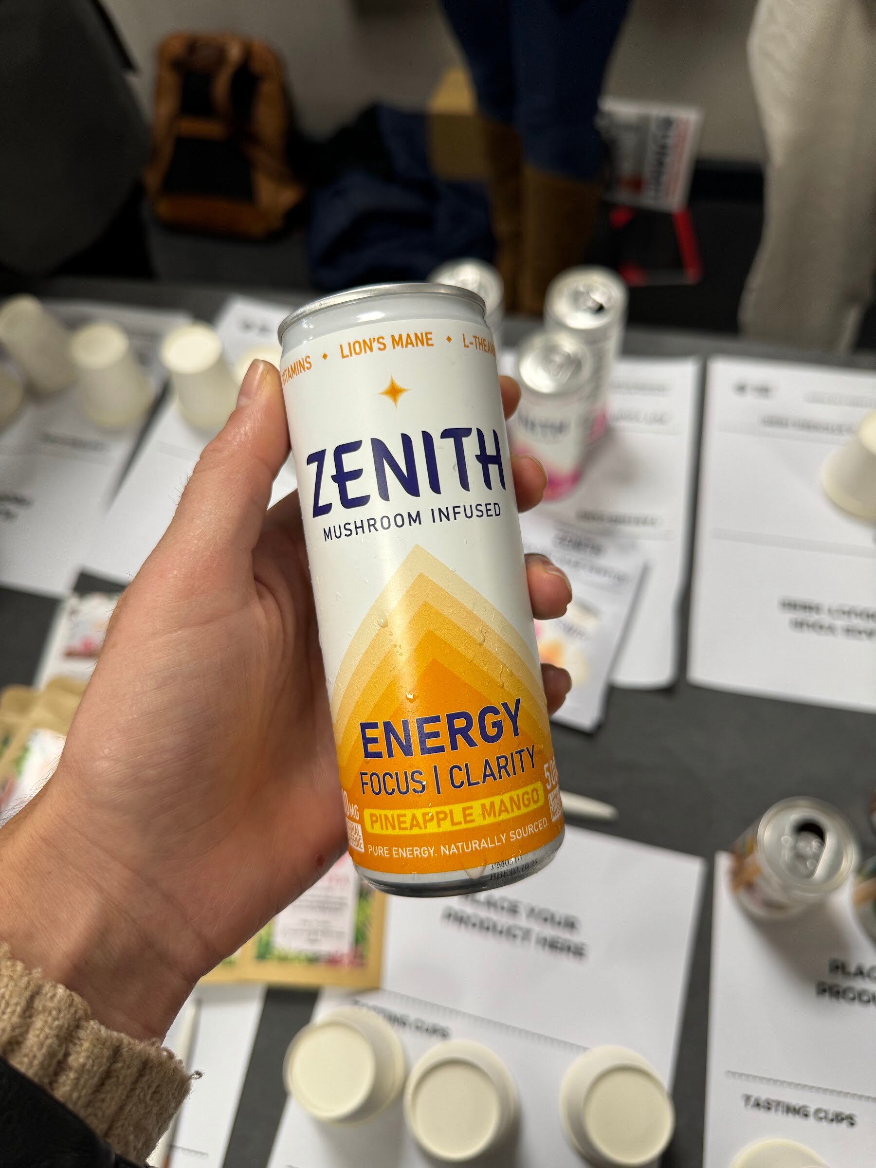 Hand holding a white and yellow can of Zenith mushroom-infused energy drink above tasting cups.
