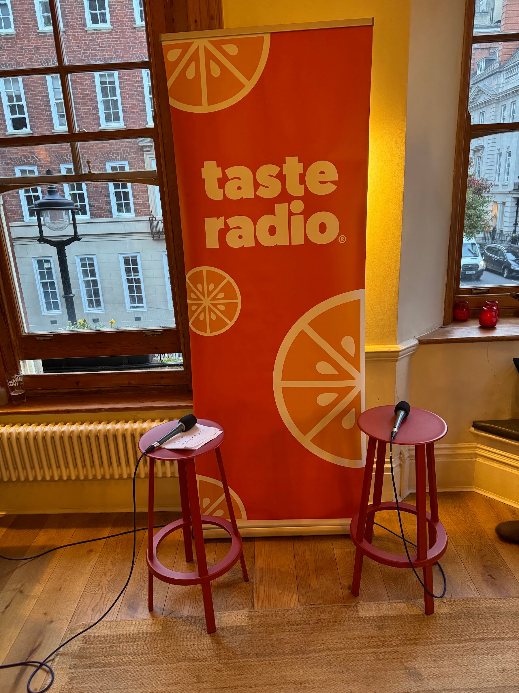 Orange Taste Radio banner beside two red stools with microphones in front of a large window.