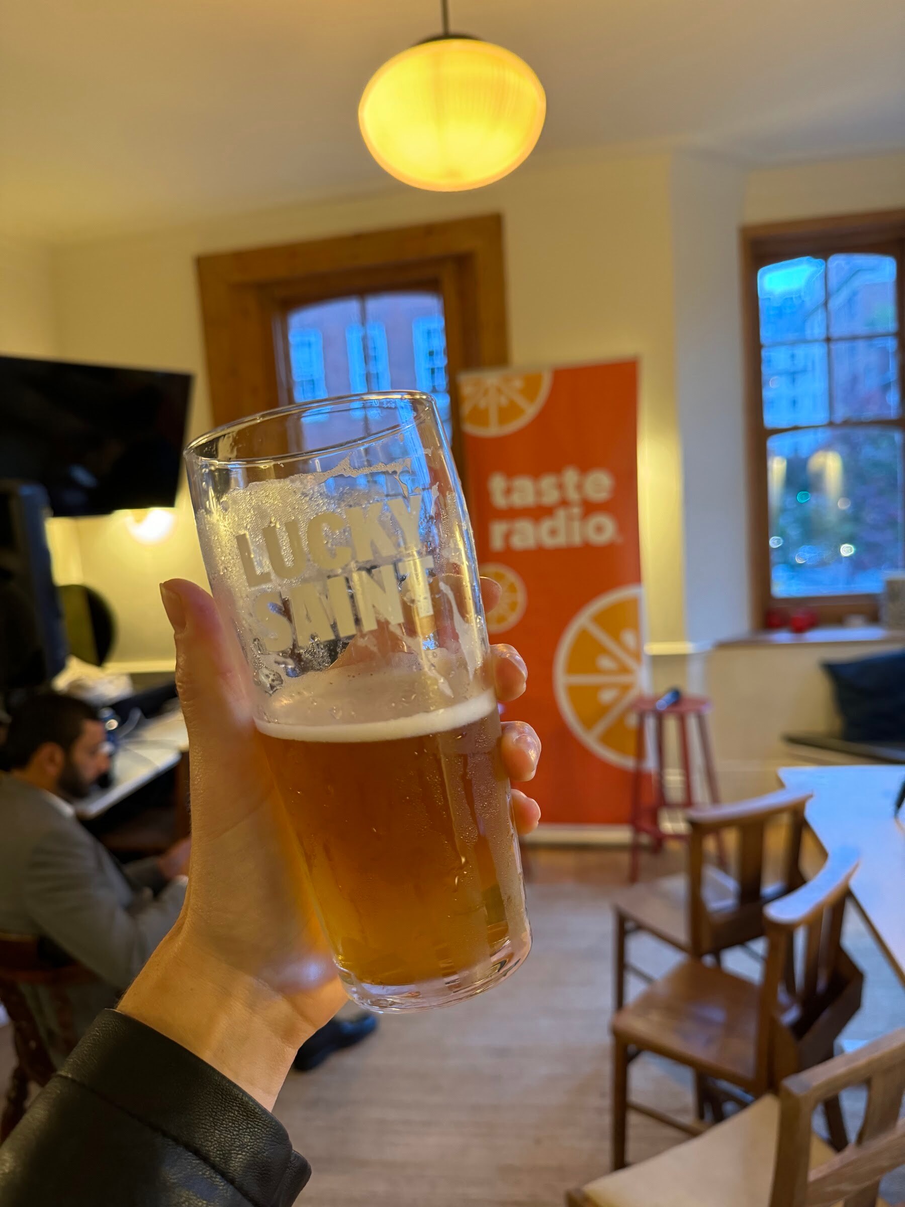 Hand holding a pint glass of beer printed with the Lucky Saint logo, with a Taste Radio banner in the background.