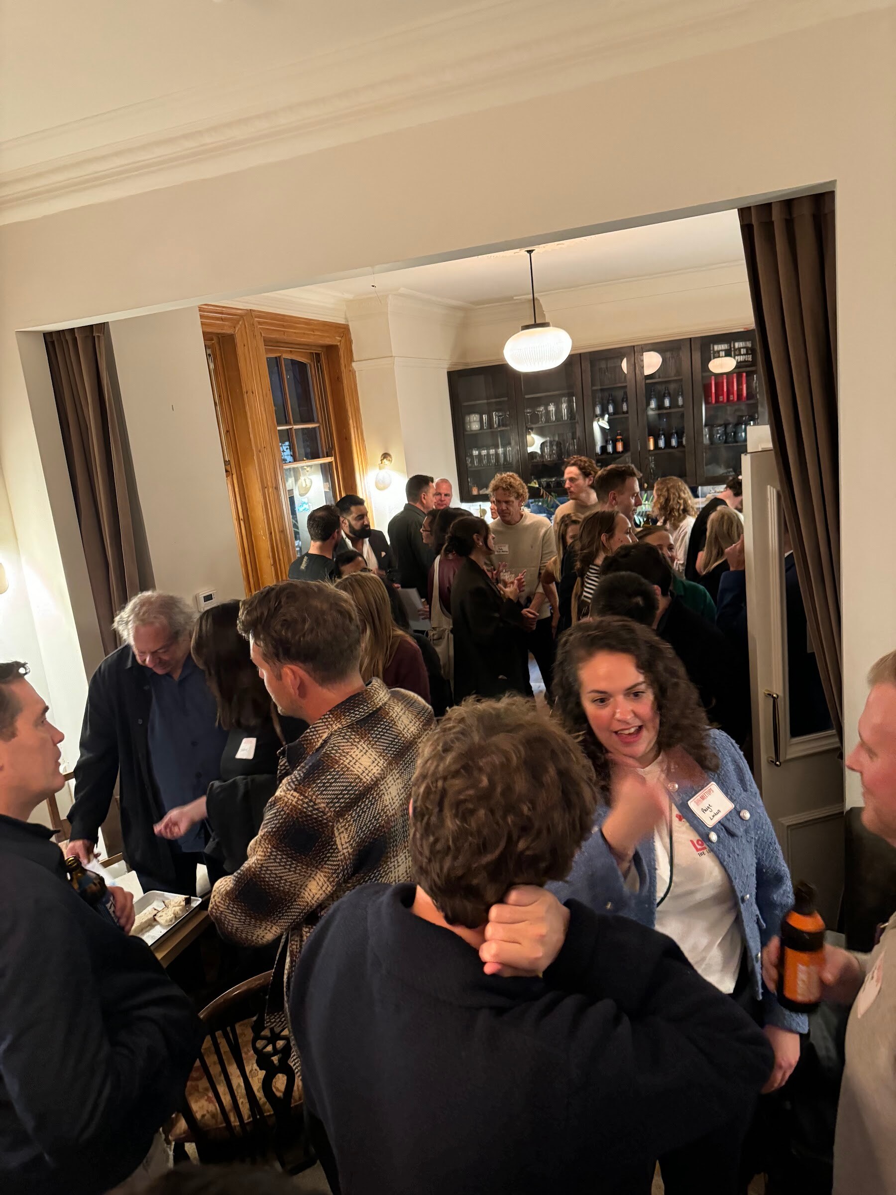 Crowd of guests talking in a warmly lit room with wood paneling and a bar area at the back.