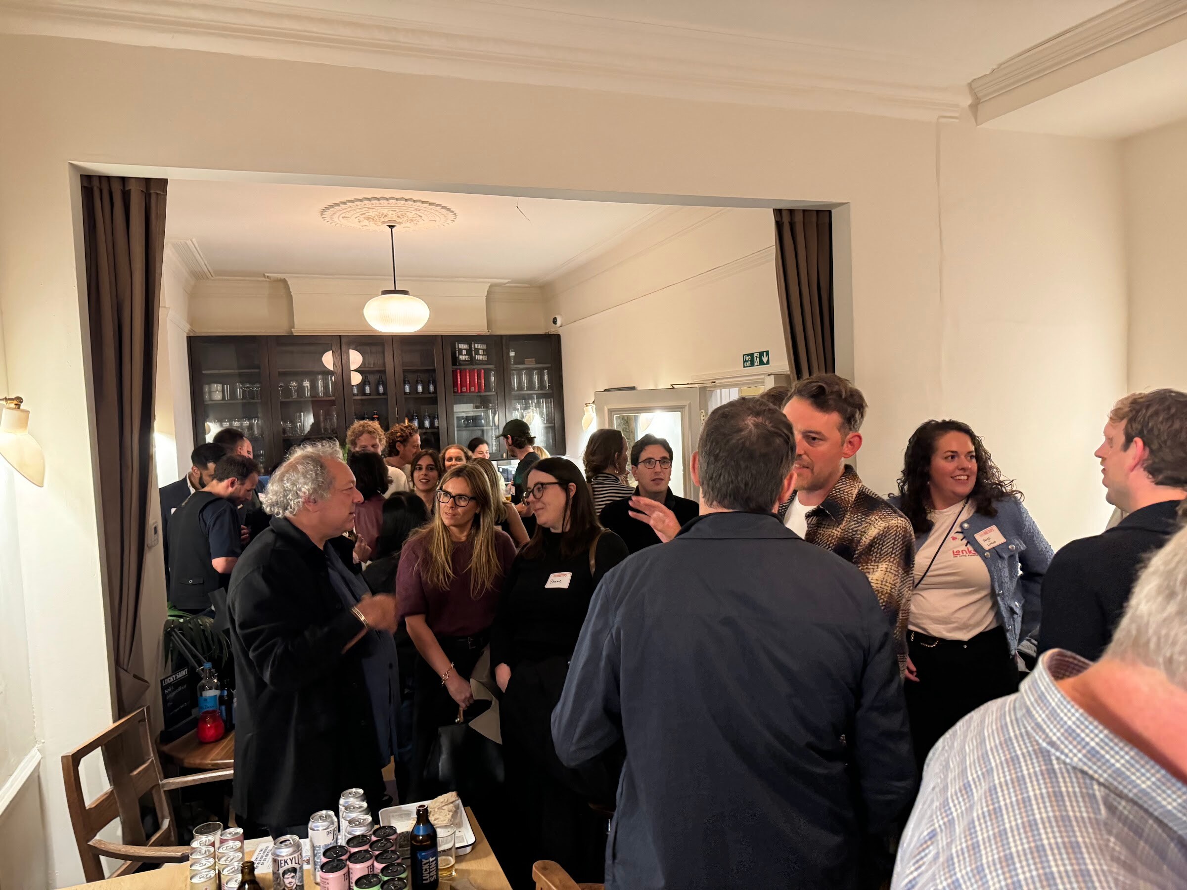 Guests talking in a warmly lit room with a table of cans and glasses in the foreground.