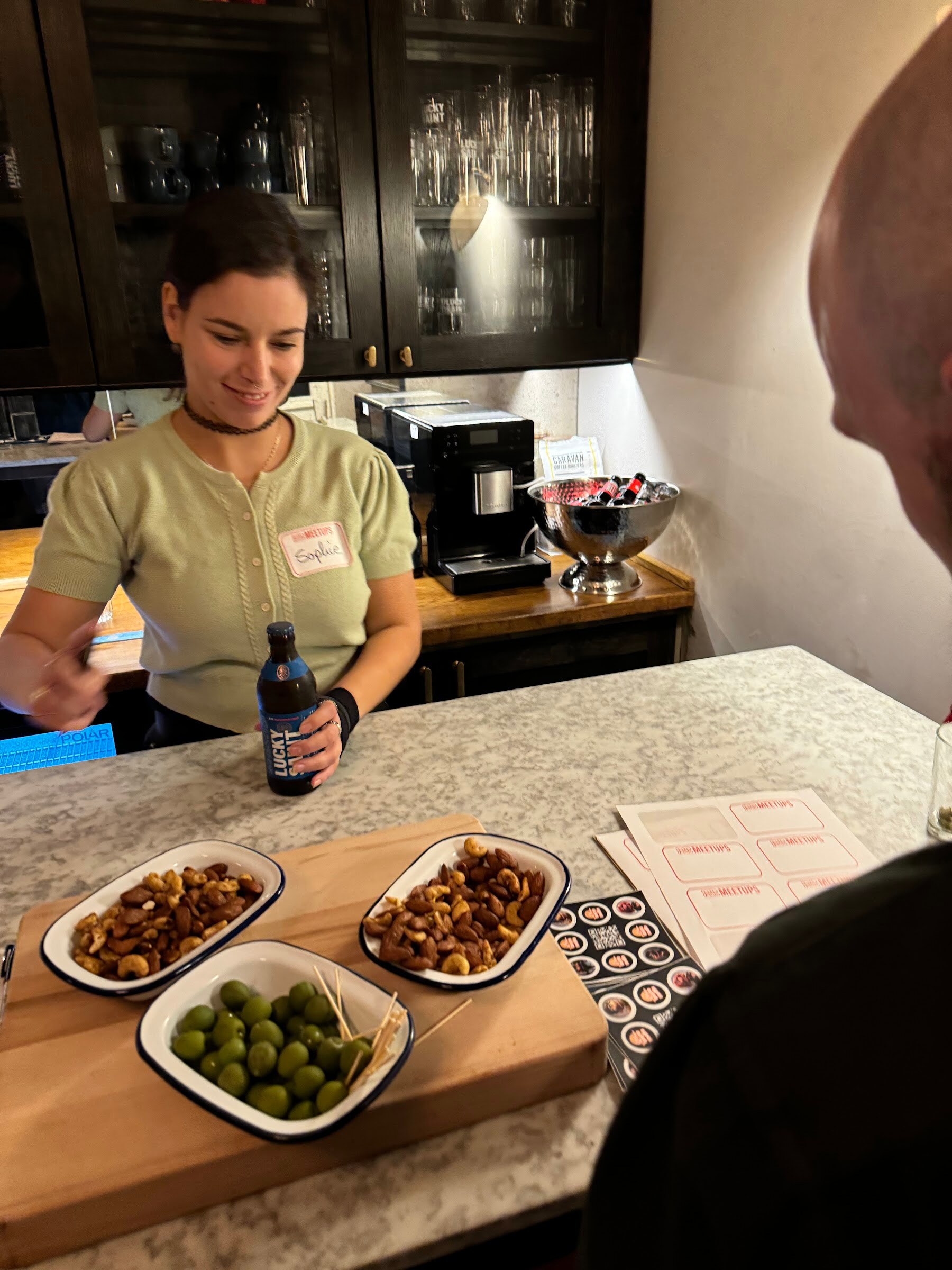 Woman in a pale green cardigan holding a dark bottle behind a marble counter with bowls of nuts and olives.