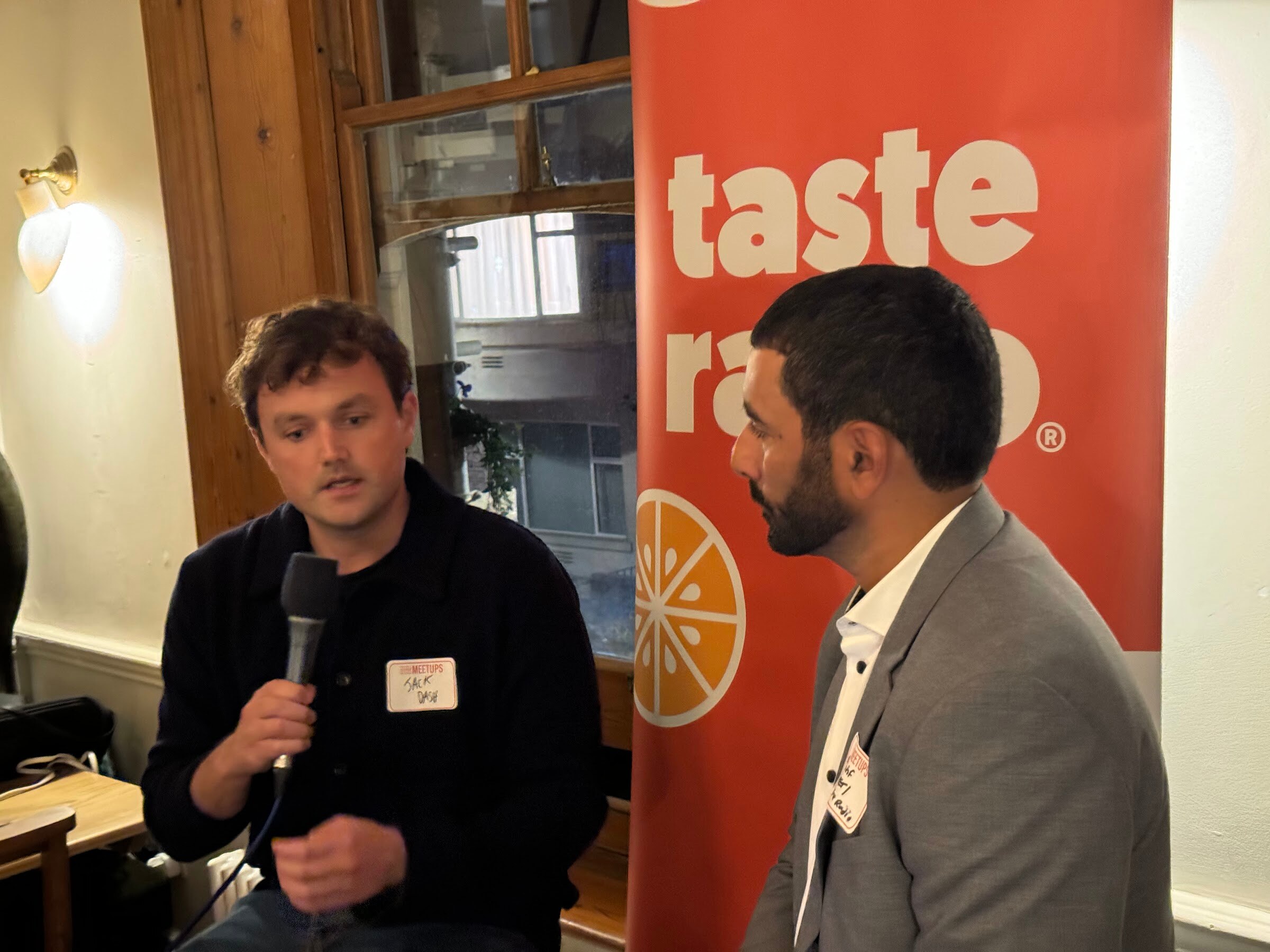 Man with a microphone and name tag speaking beside the Taste Radio interviewer in front of an orange Taste Radio banner.
