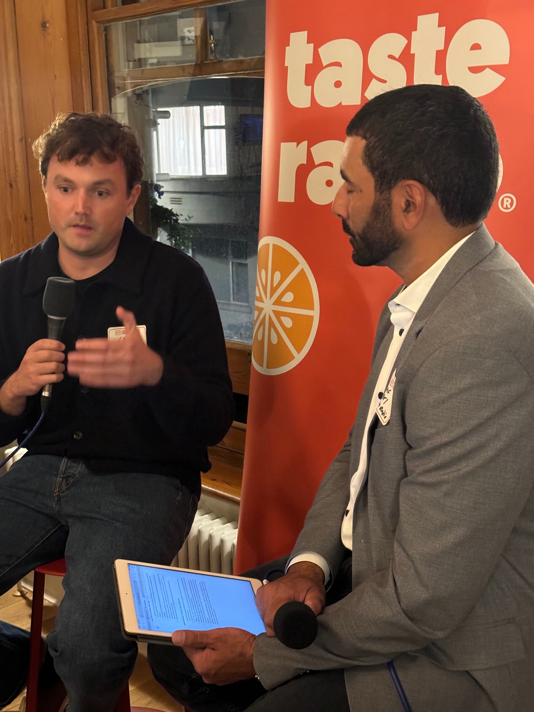 Seated guest with a microphone gesturing, beside the interviewer holding a tablet, in front of a Taste Radio banner.