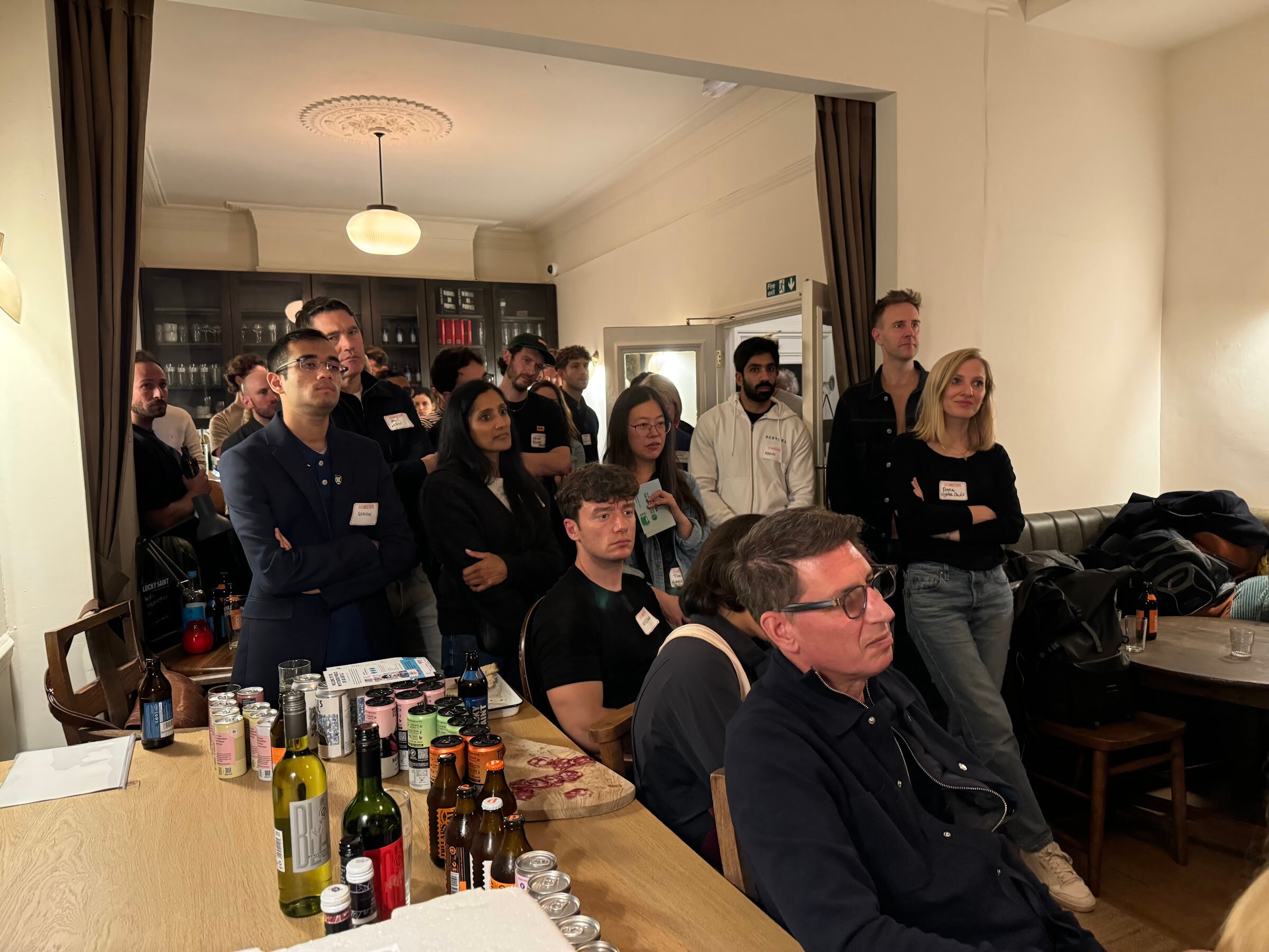 Standing audience at the back of the room listening to the interview, with cans and bottles on a table in the foreground.