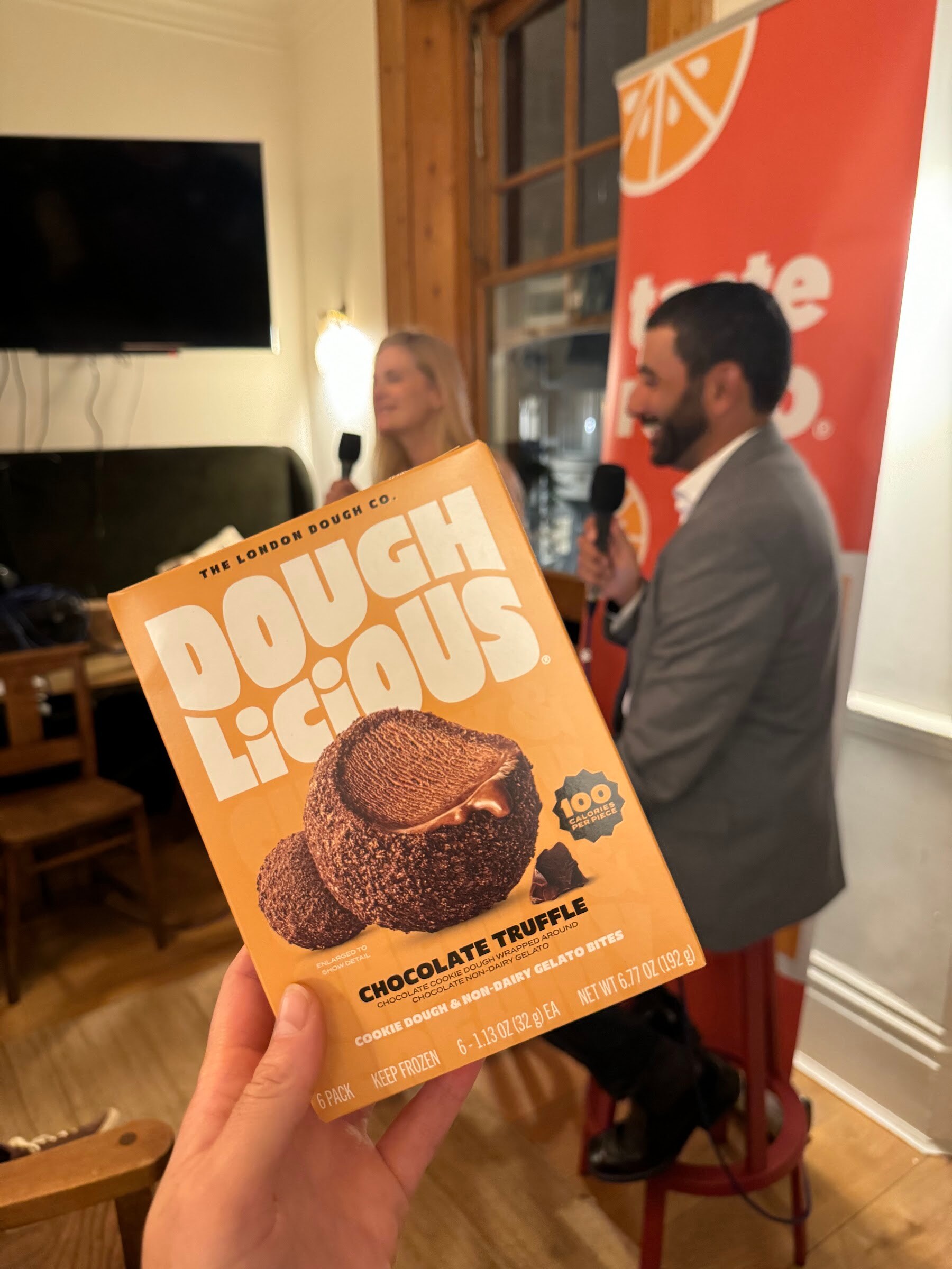 Hand holding a Doughlicious Chocolate Truffle box in the foreground, with two people being interviewed in the background.
